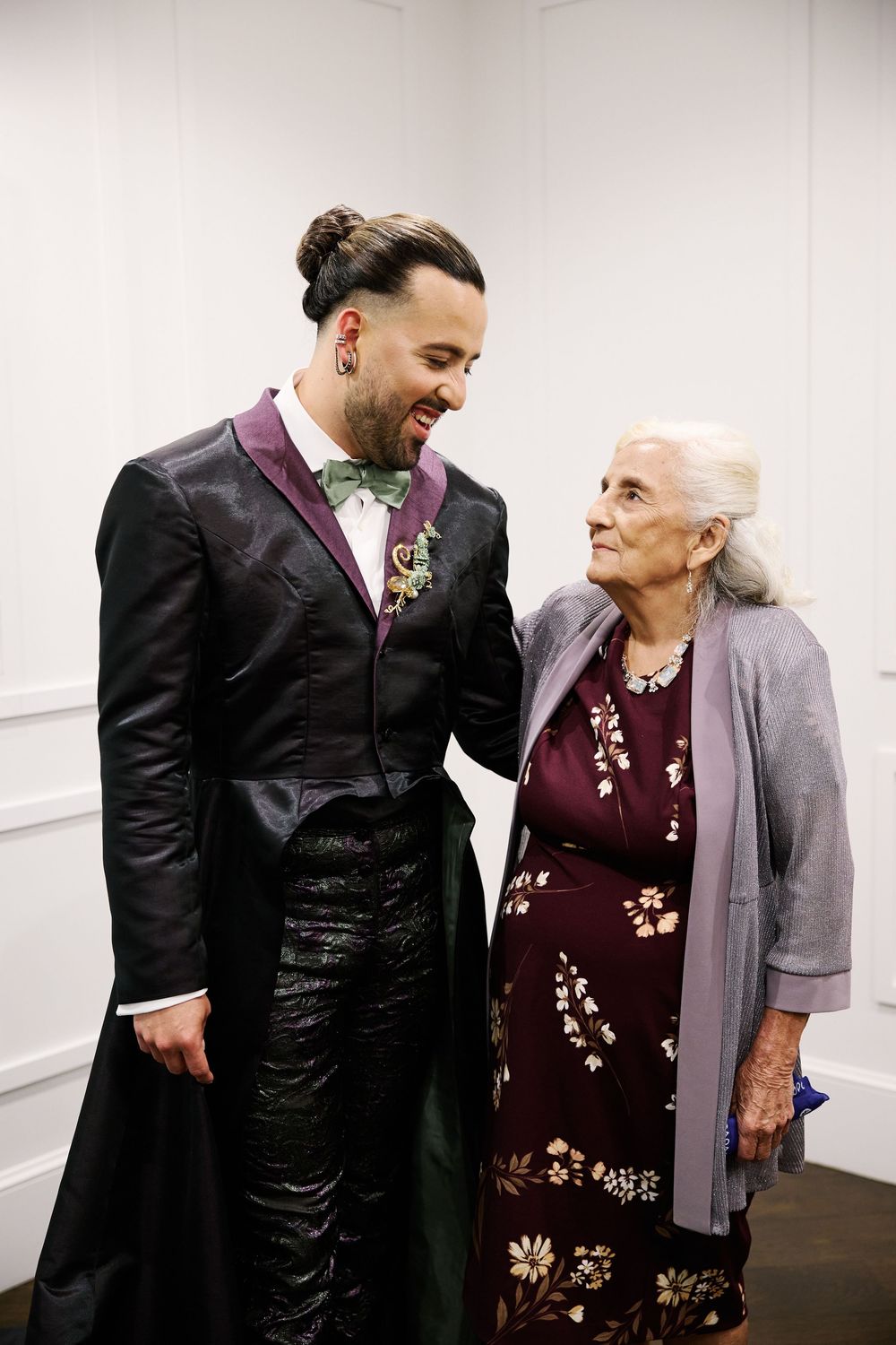 A groom in a black tuxedo with green bow tie shares a joyful moment with an elderly woman at a wedding.