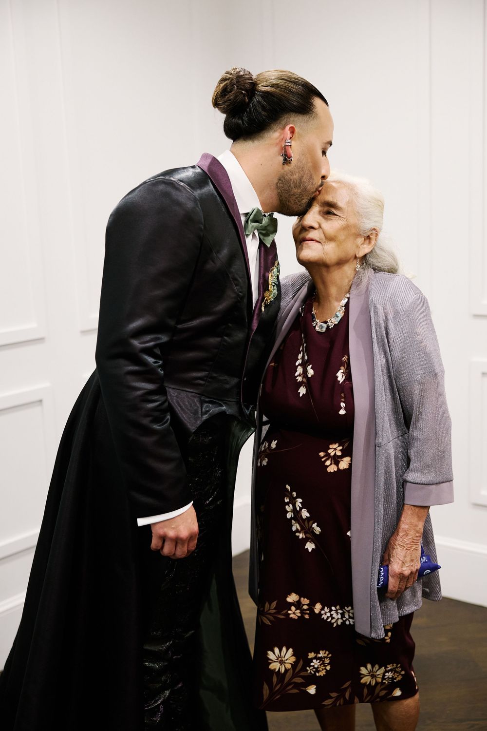 Groom in black leather wedding attire kisses elderly woman on cheek at wedding celebration.