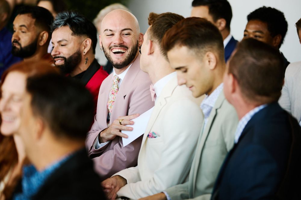 Group of smiling wedding guests in formal attire laughing and socializing during a ceremony.