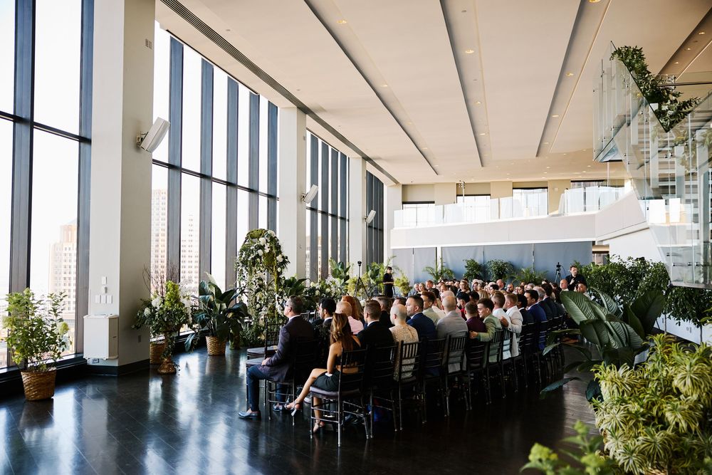 Guests seated at a modern indoor wedding reception venue with floor-to-ceiling windows and lush greenery.
