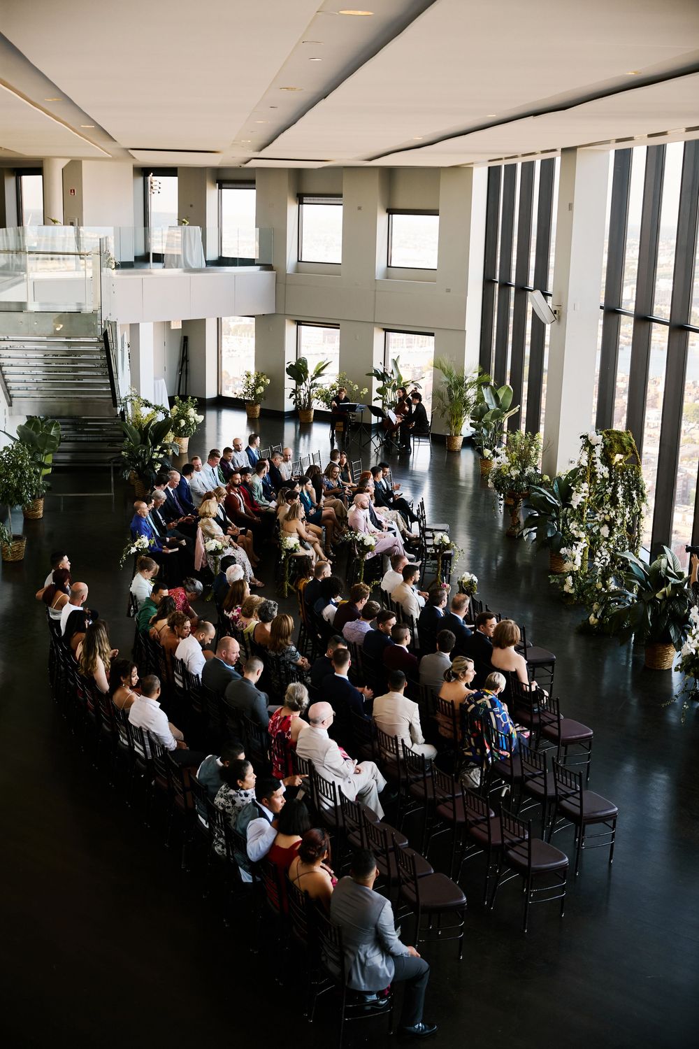Overhead view of wedding guests seated in modern venue with large windows and tropical plants.
