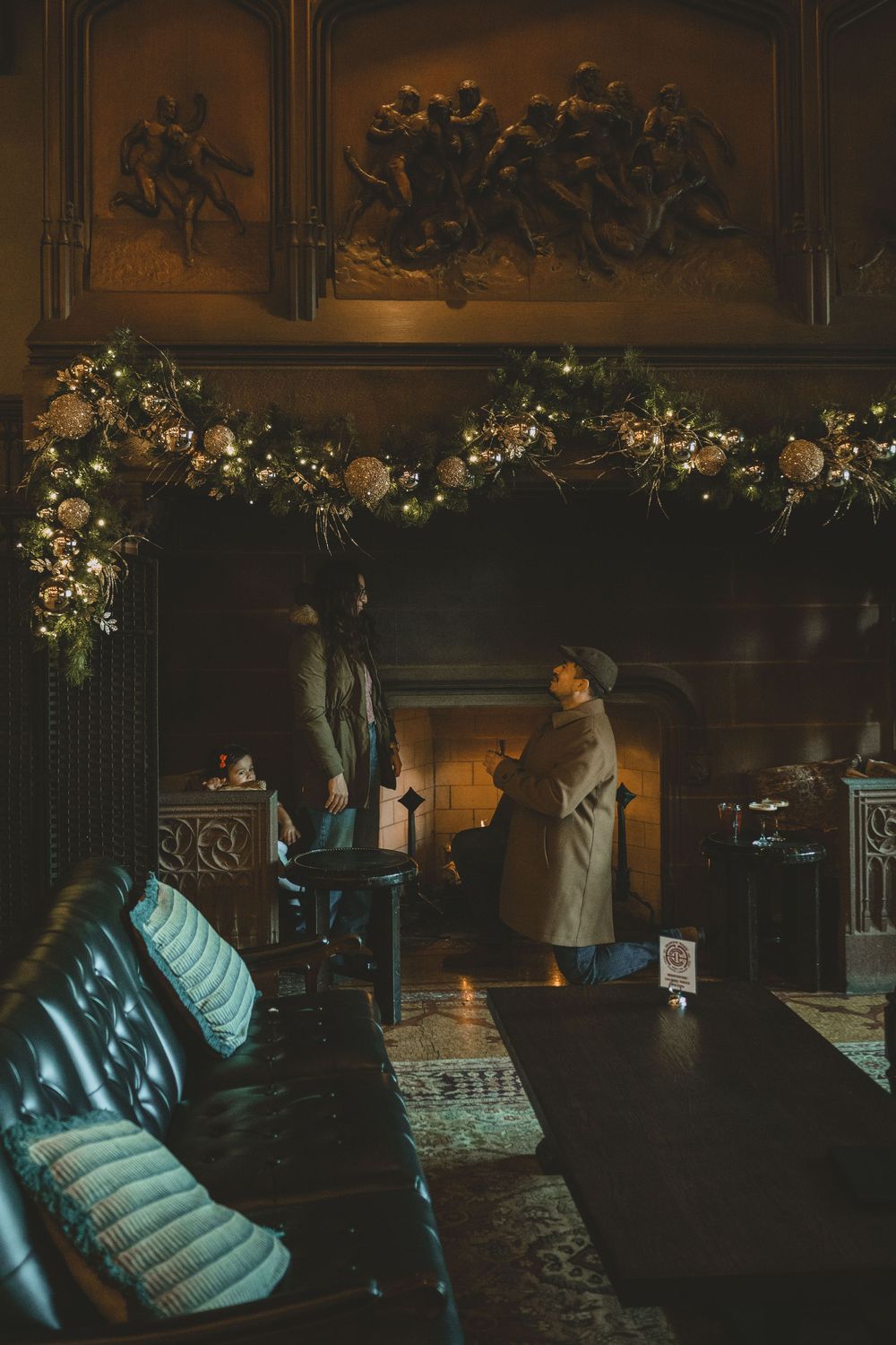 Elegant dark interior with Christmas garland over fireplace, two people standing in warm ambient light near leather sofa.