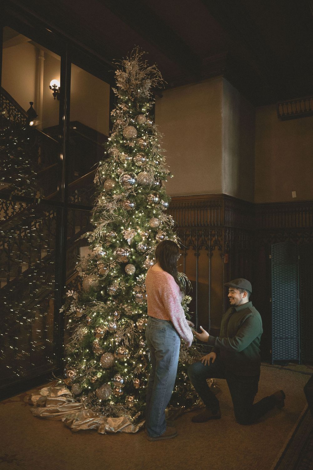 Man kneeling in marriage proposal before decorated Christmas tree in elegant wood-paneled interior.