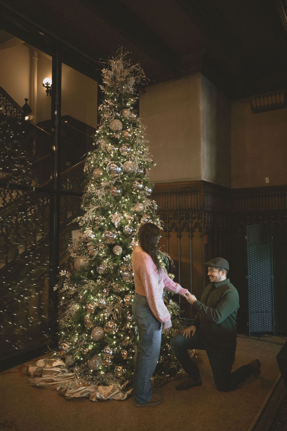 Man proposes to woman on one knee beside a decorated Christmas tree in an elegant wood-paneled room.