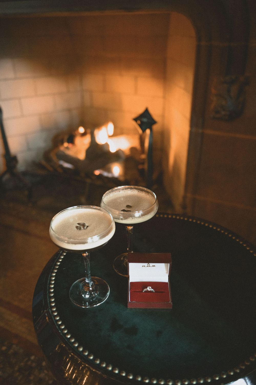 Two champagne coupes and an engagement ring in a red box on a dark table beside a warm glowing fireplace.