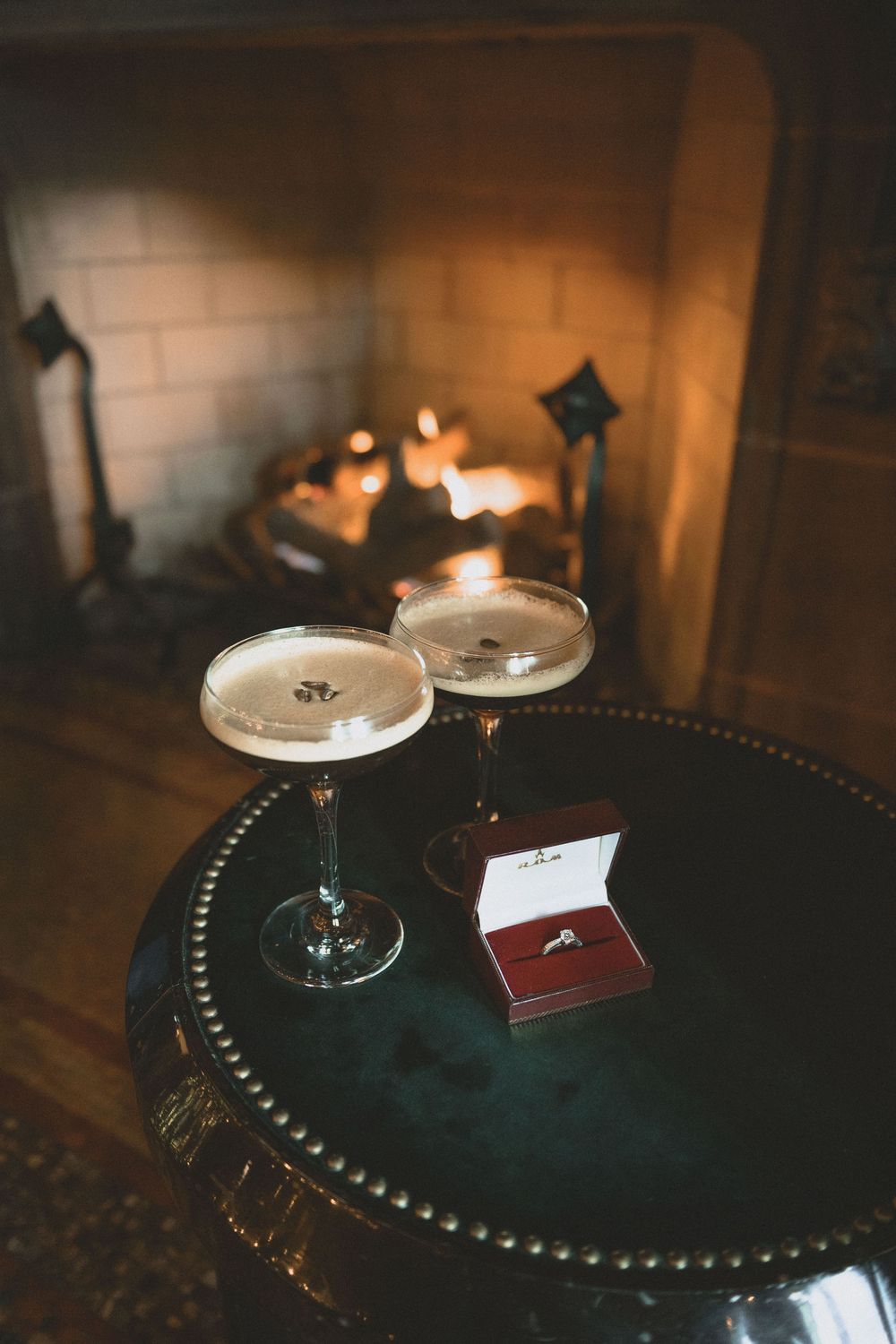 Two champagne coupes and a diamond engagement ring in a red box on a dark table beside a glowing fireplace.