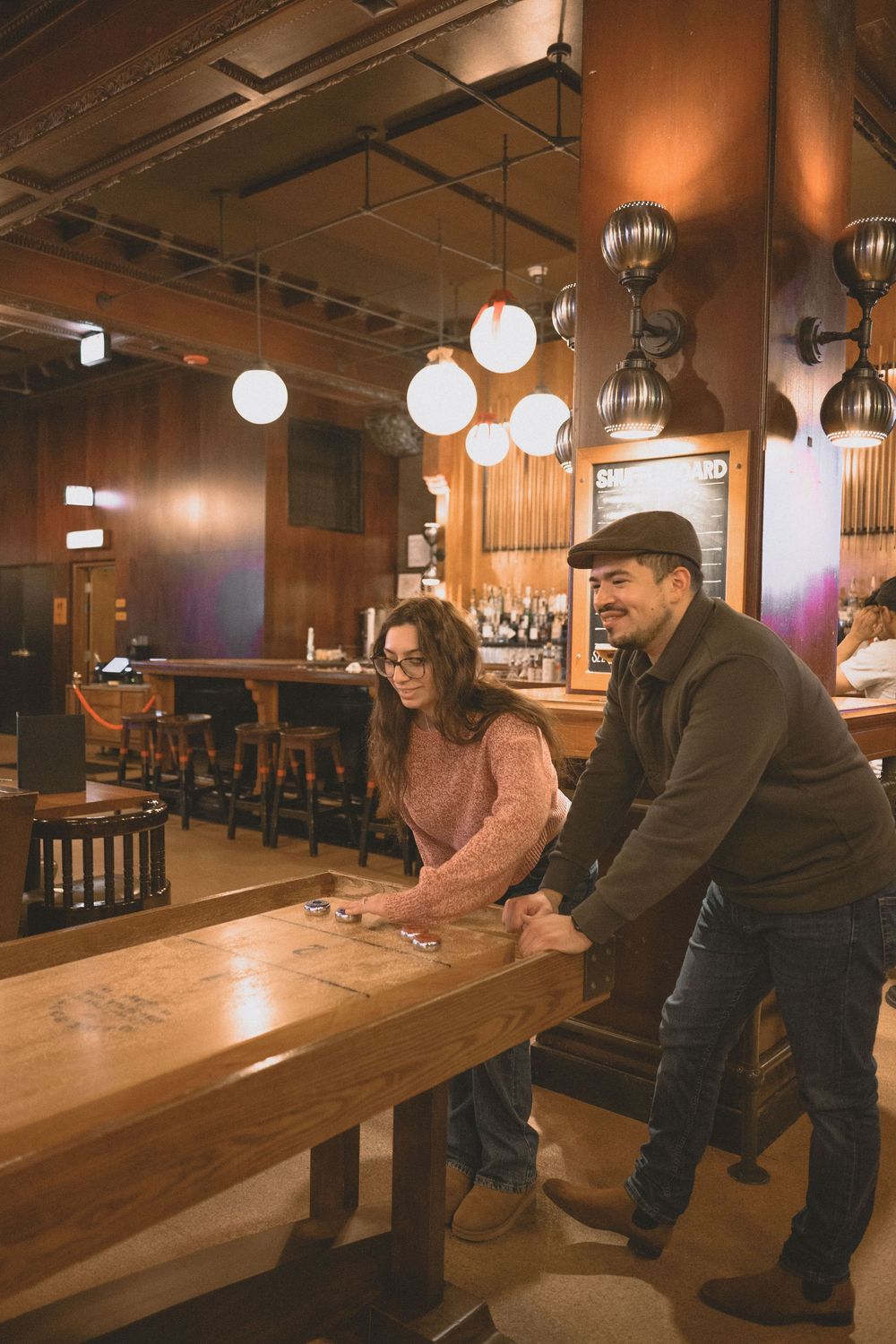 A couple plays shuffleboard at a cozy bar with warm lighting and wooden decor.