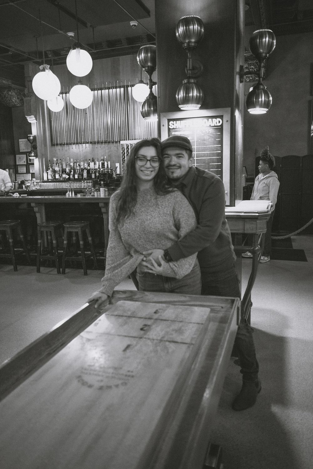 Black and white photo of a smiling couple posing together at a shuffleboard table inside a bar restaurant.