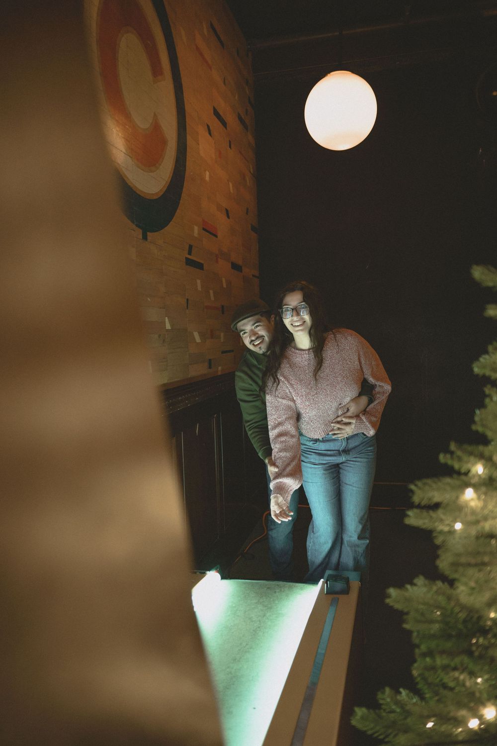 Couple holding hands and smiling near a Christmas tree in a warmly lit brick interior at night.