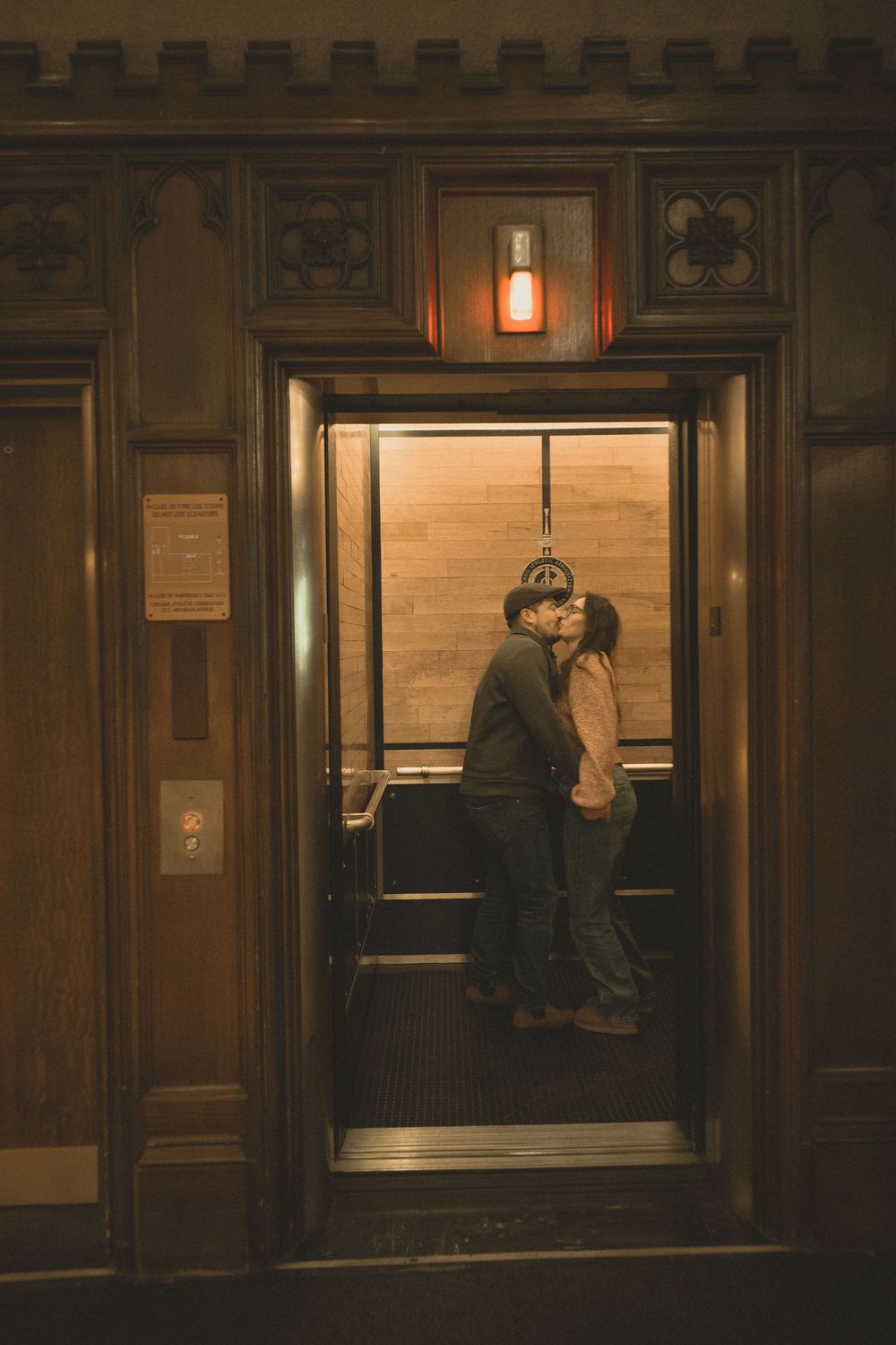 Couple sharing a romantic kiss inside a vintage wooden elevator with warm amber lighting.