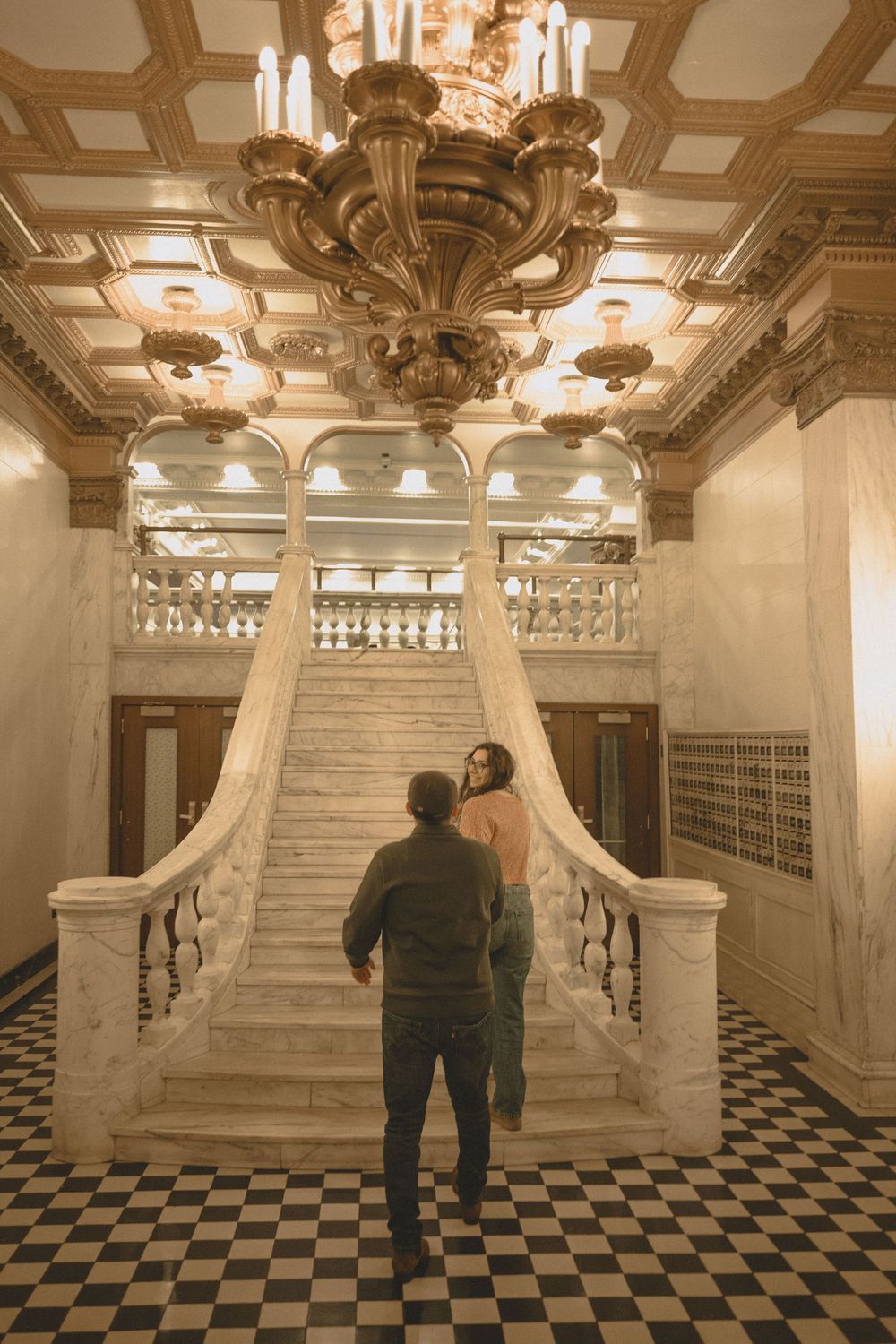 Couple standing before grand marble staircase in ornate ballroom with chandelier and checkered floor.