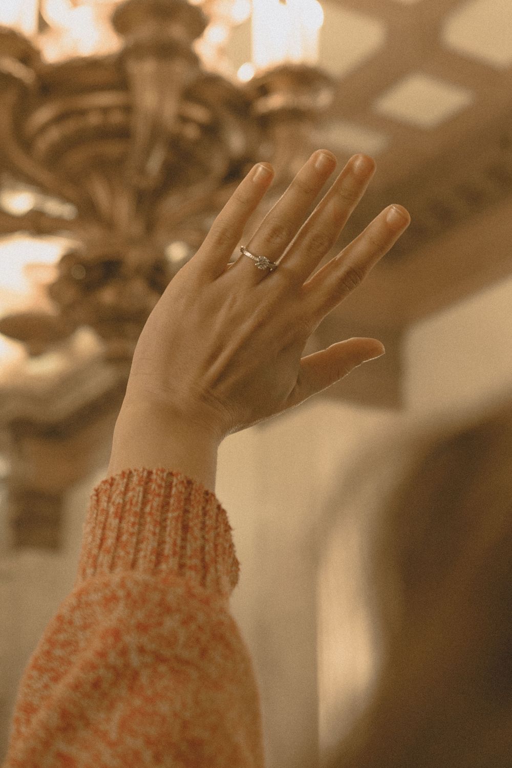 A hand wearing a delicate silver ring raised up, with an ornate chandelier in the background, wearing an orange knit sweater.