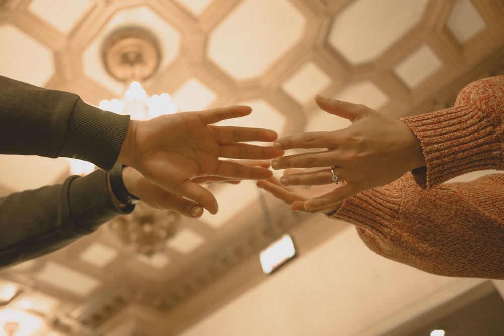 Two people reaching out to touch hands, wearing casual clothing, with an ornate ceiling visible in the background.