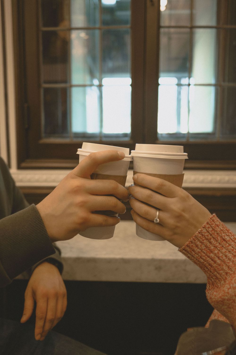 Two people toasting with takeaway coffee cups near a window, one wearing a ring on their finger.