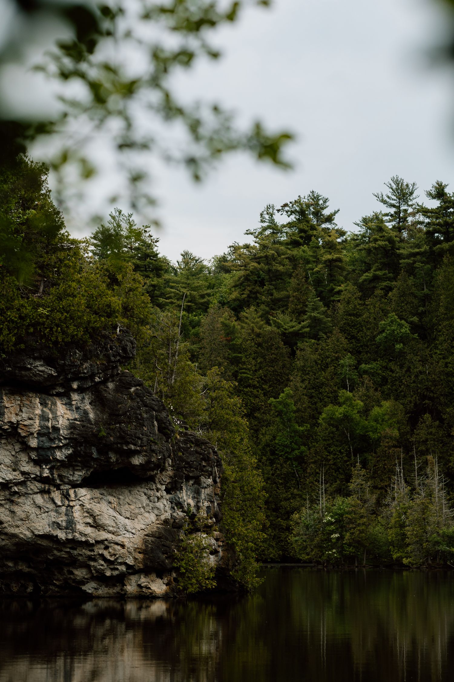 Rocky cliff beside a calm forest lake surrounded by dense green pine trees reflected in still water.