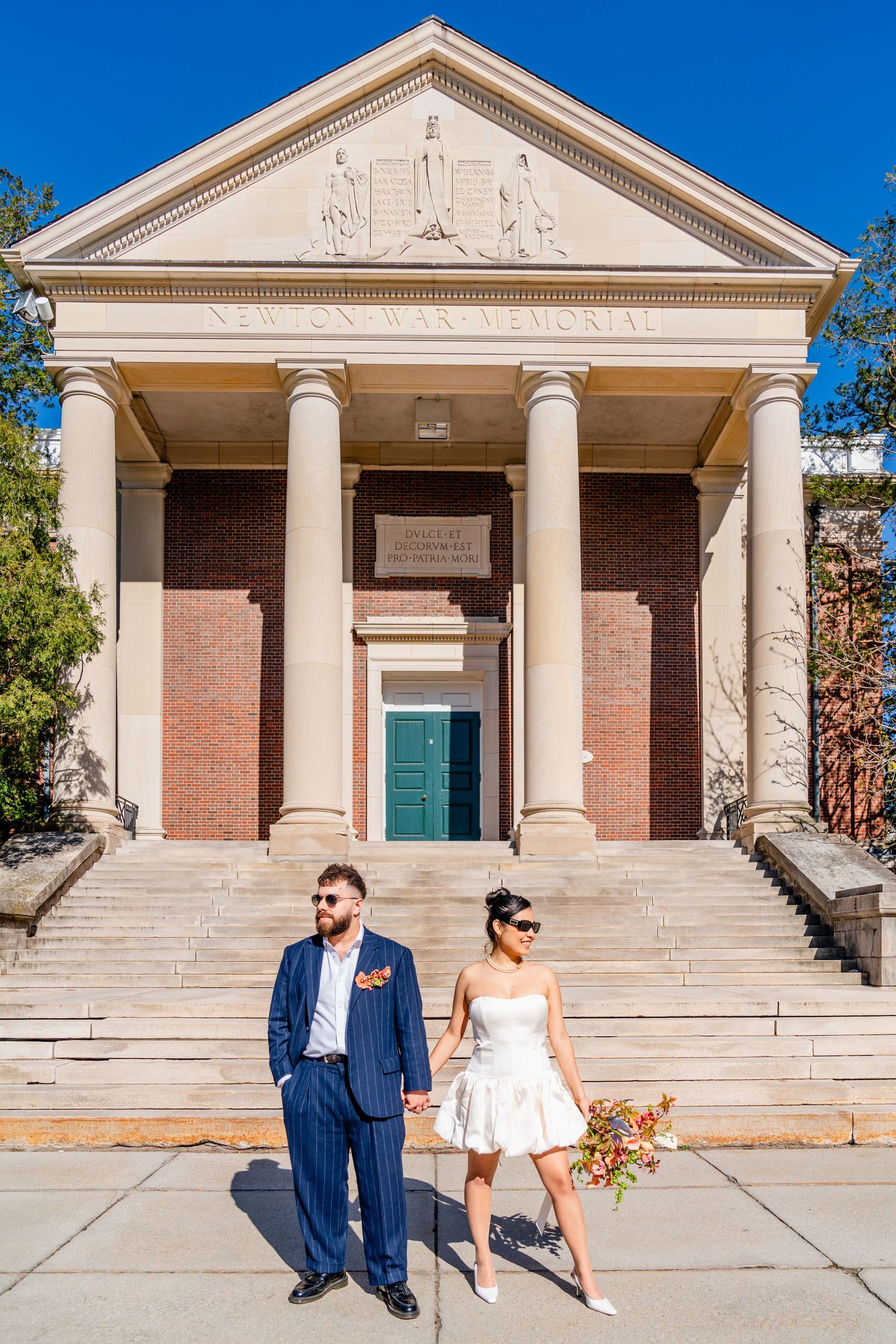 Couple in wedding attire holding hands on steps of Newton War Memorial building on sunny day.