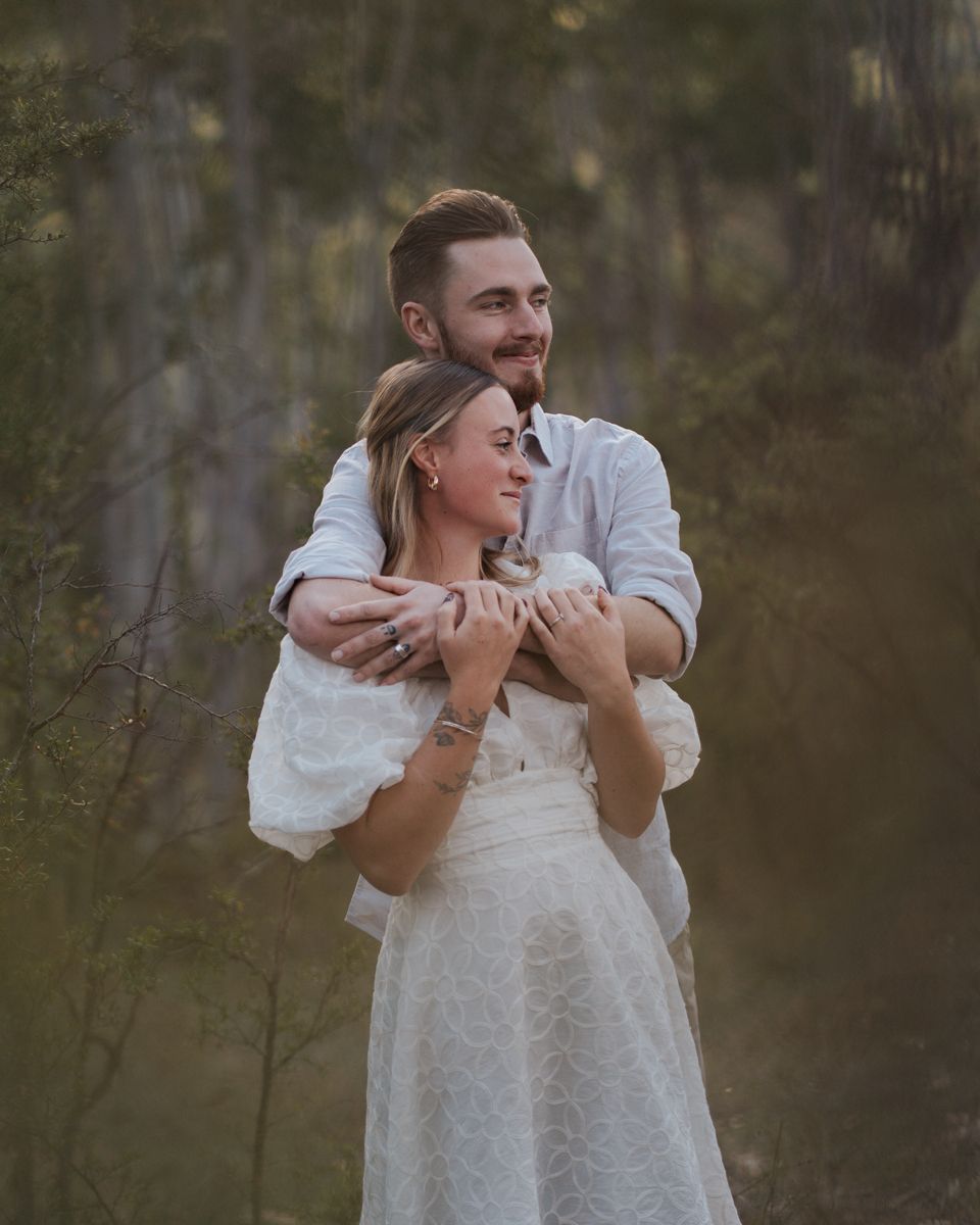Couple outdoors in white outfits, man hugging woman from behind, smiling in a soft forest setting in rouse hill engagement photoshoot