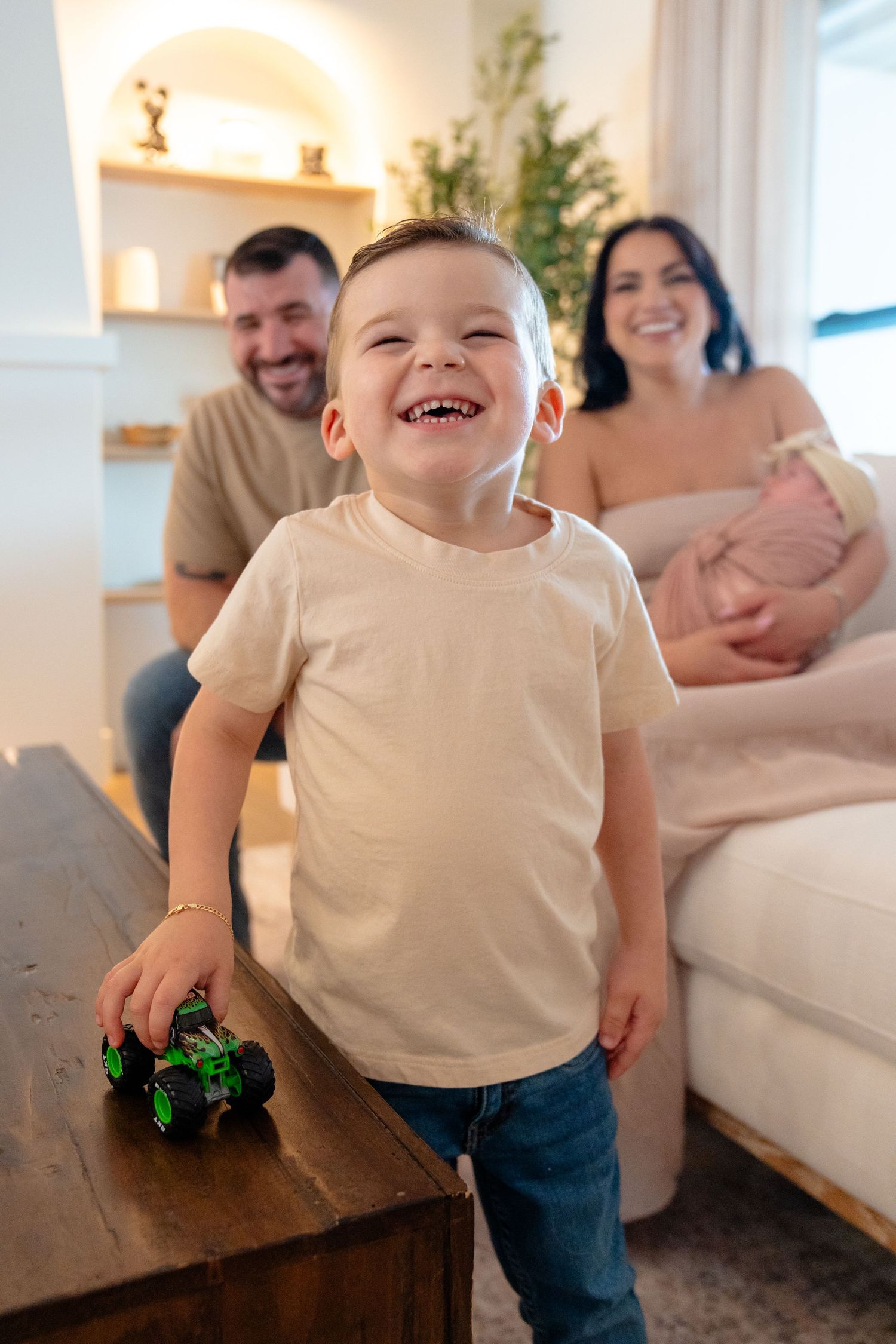 Happy toddler boy laughing while playing with toy car, parents smiling in background with newborn baby.