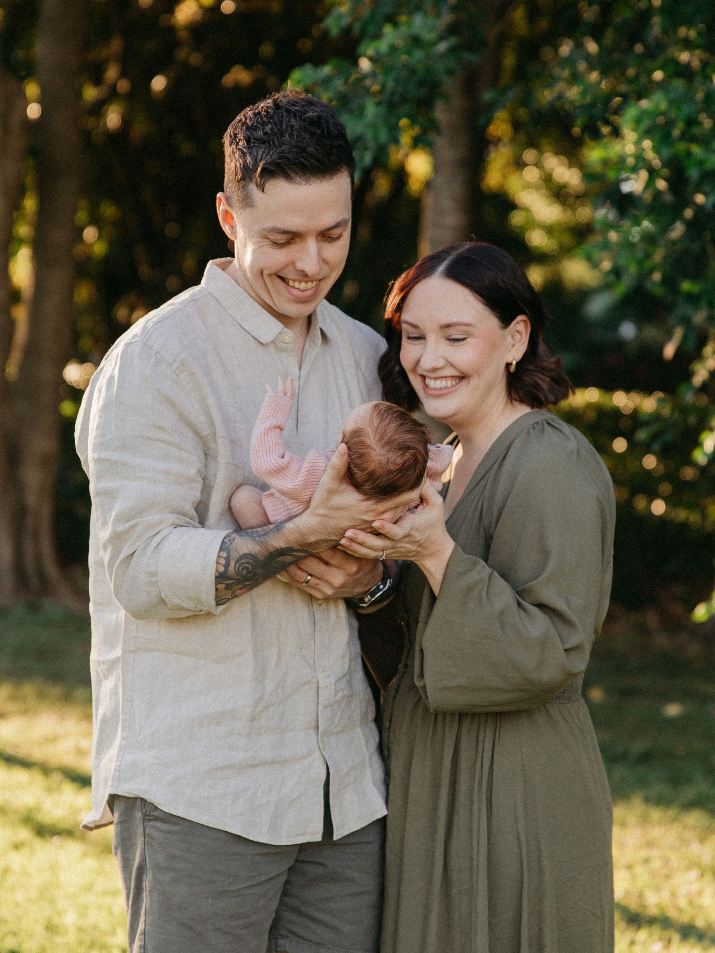 Happy couple smiling at their newborn baby outdoors in a sunlit garden setting.