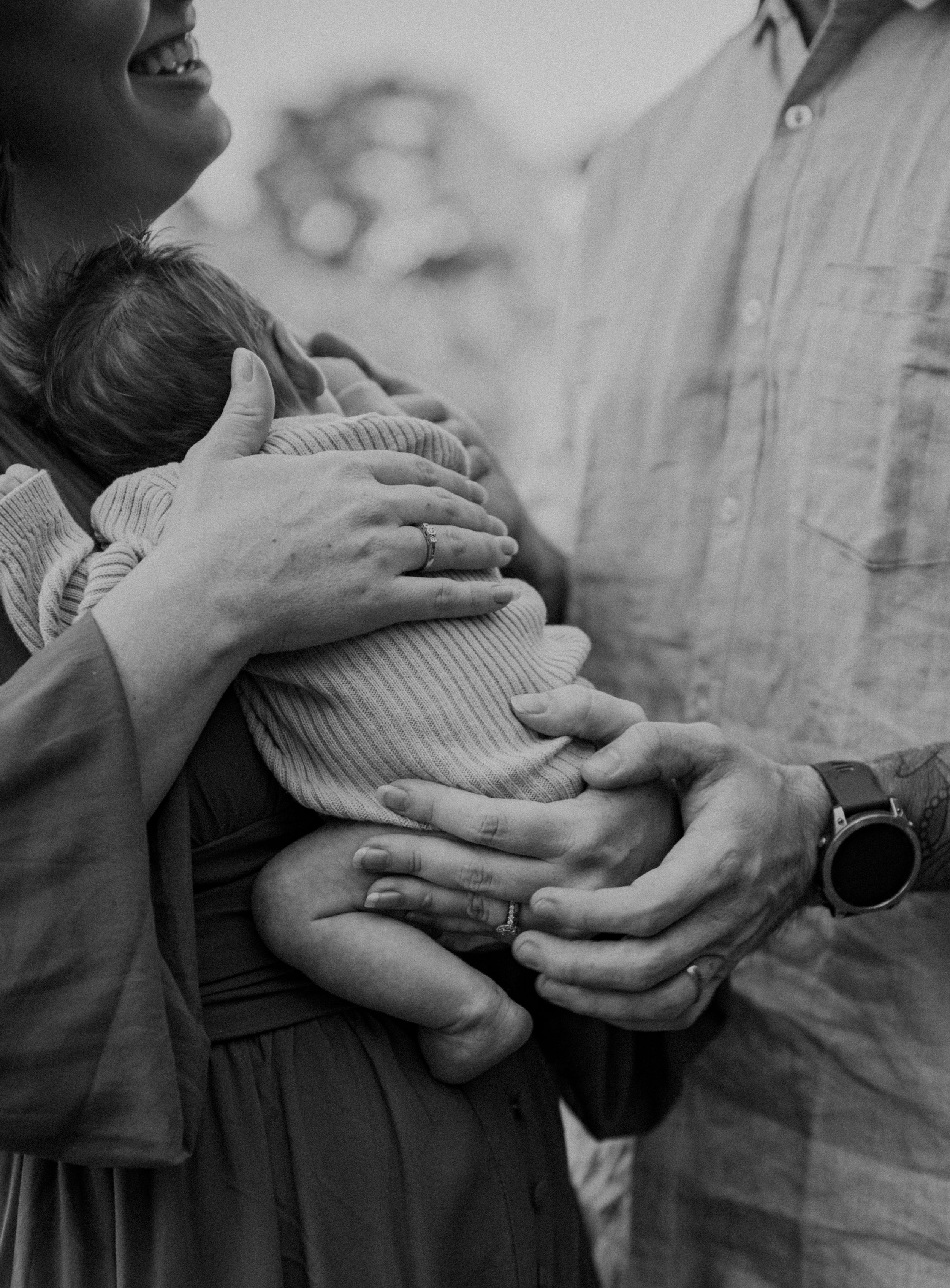 Black and white photo of parents cradling their newborn baby, both wearing wedding rings, in an outdoor setting.