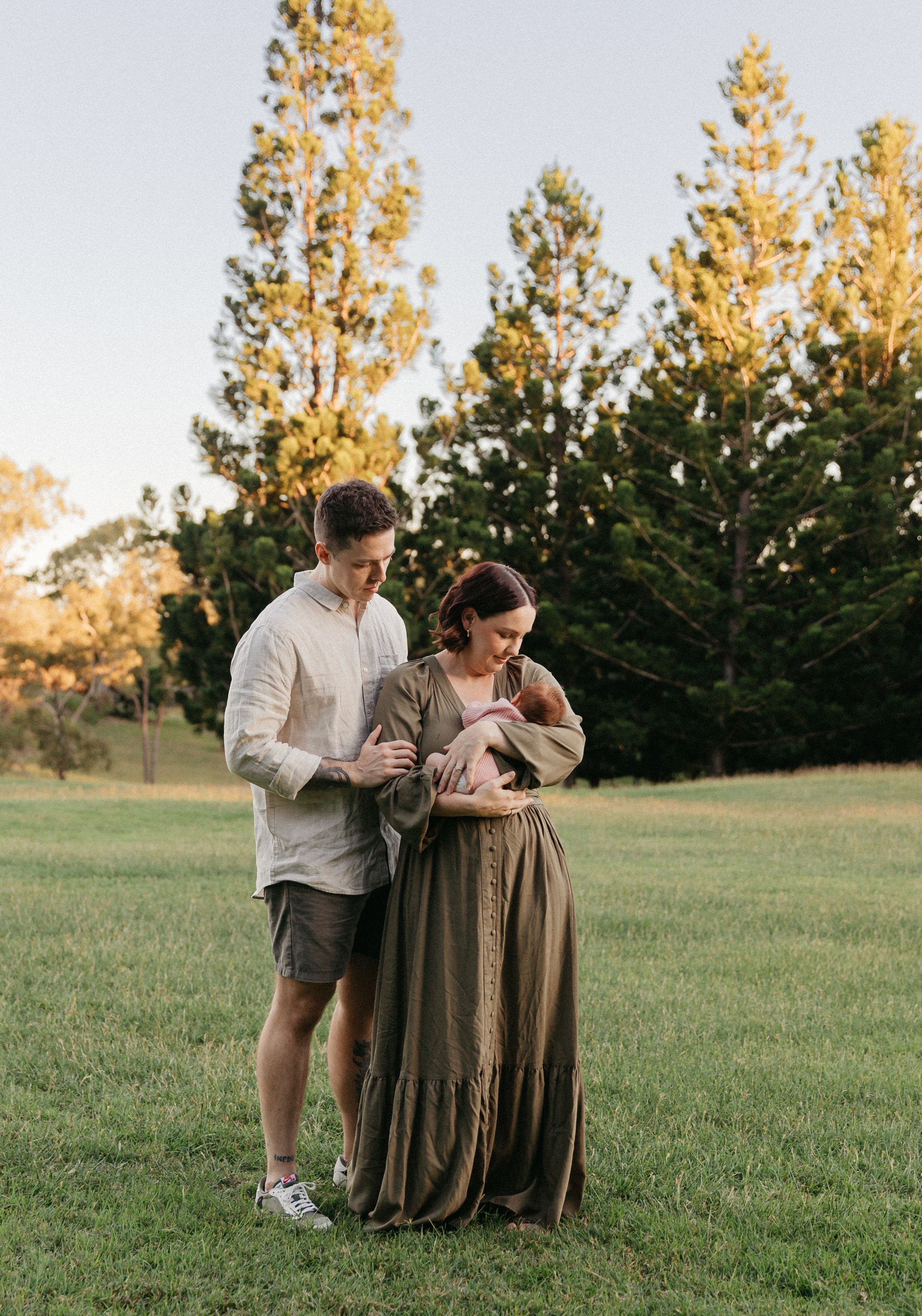 A couple stands in a sunlit green field holding their newborn baby, surrounded by tall trees at golden hour.