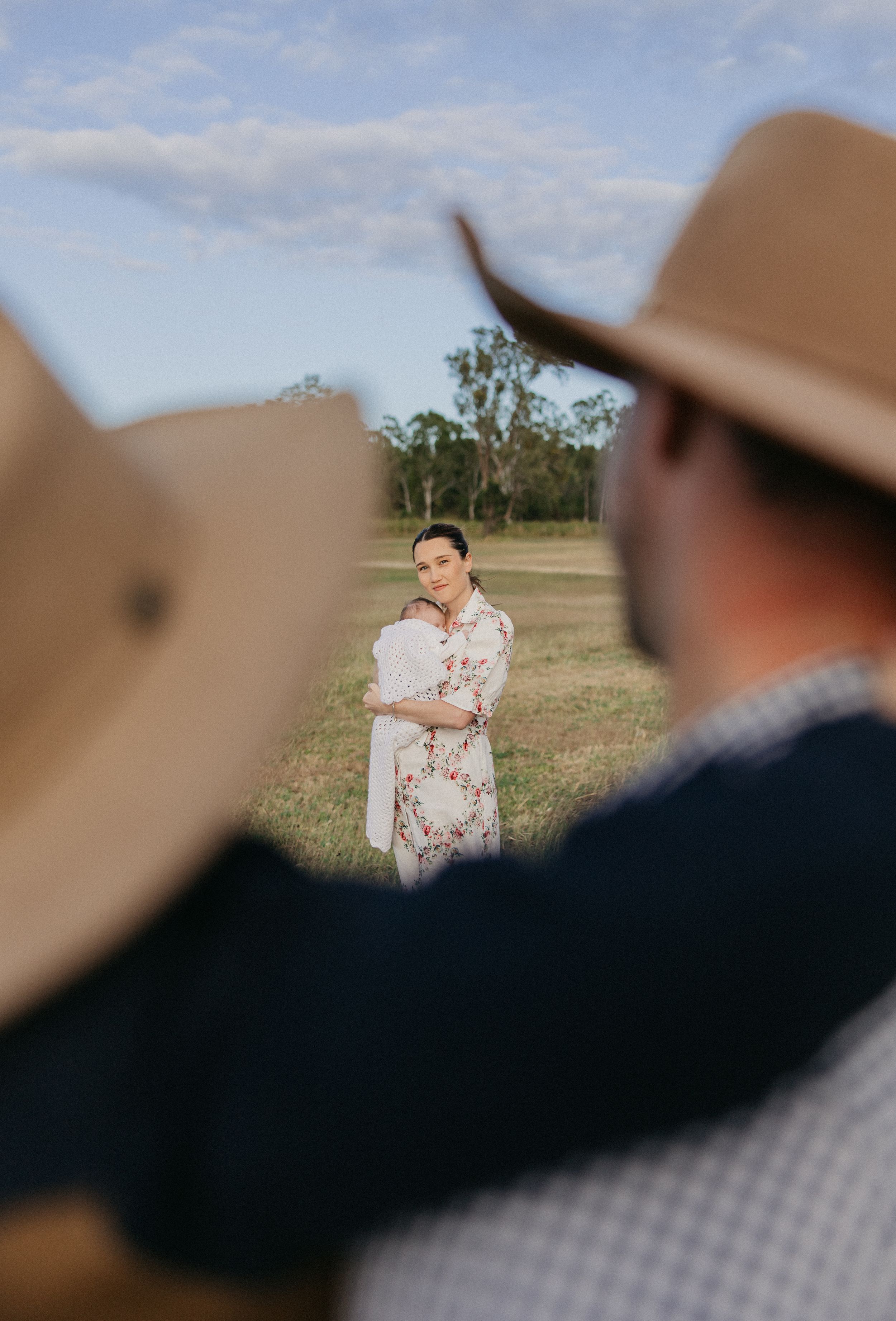 A woman in a floral dress holds a baby in a green field, framed by two people in cowboy hats in the foreground.