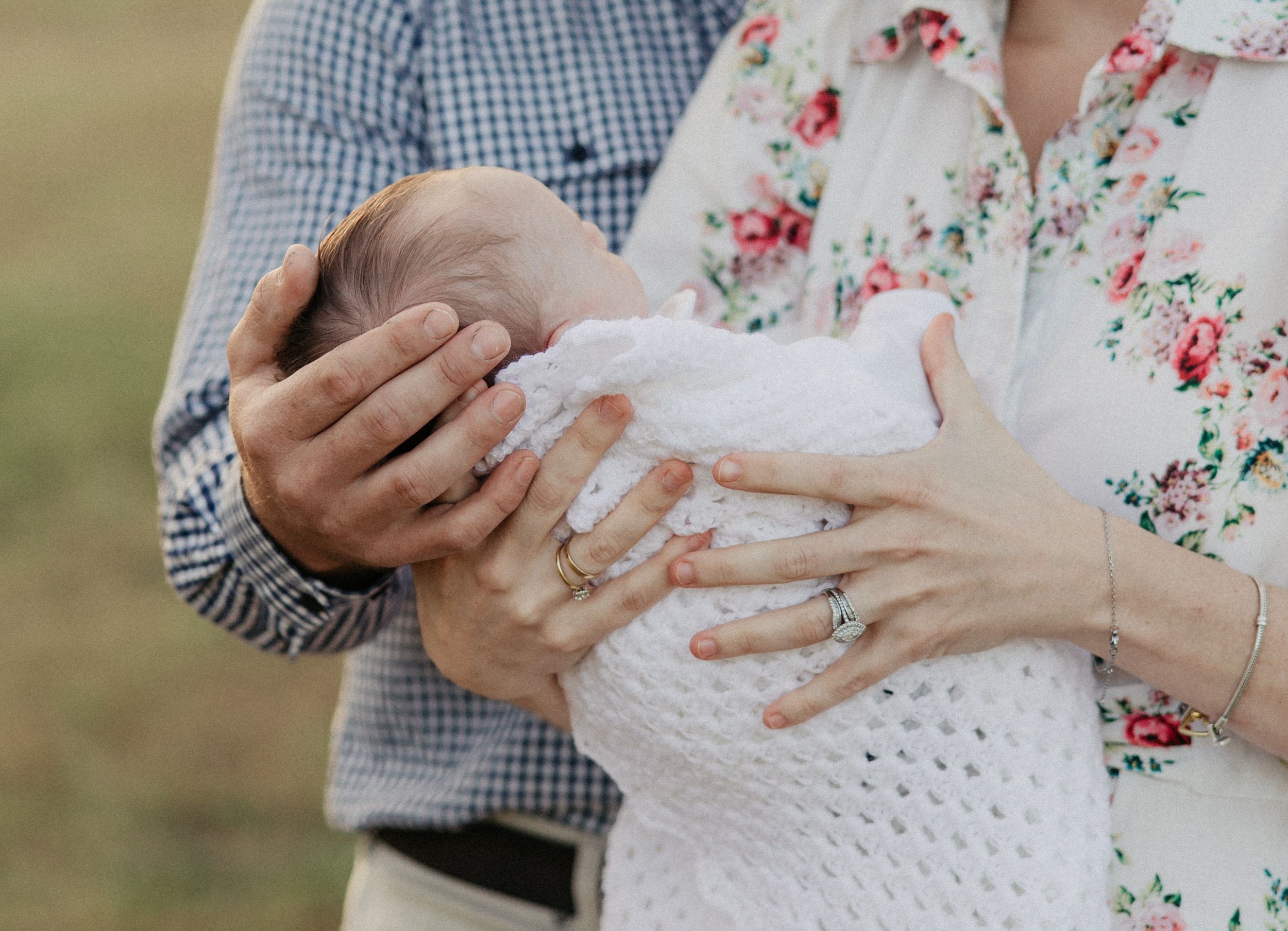 Parents cradling a newborn baby in their hands outdoors, mother wearing floral blouse and rings.