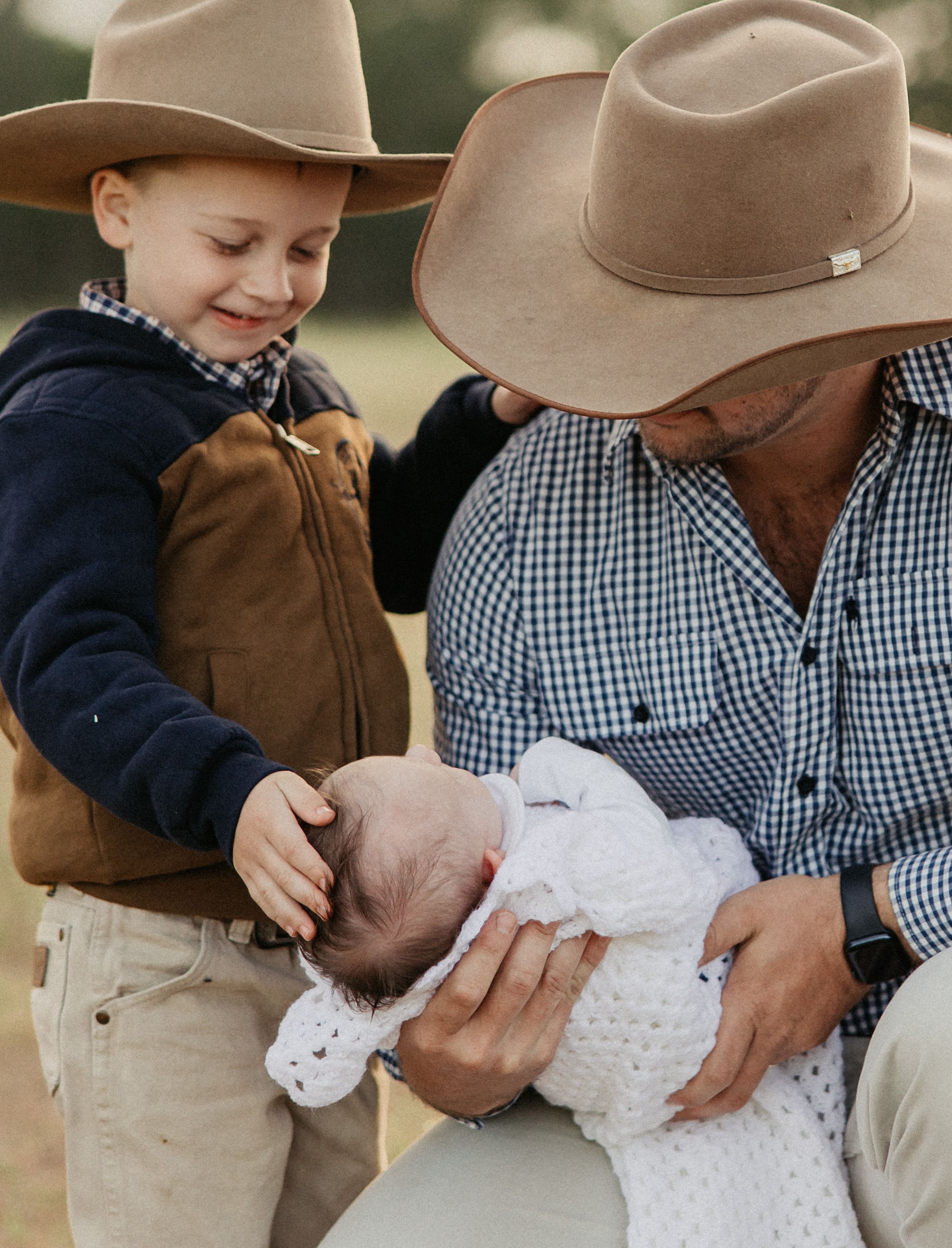 A man in a cowboy hat holds a newborn baby while a young boy in a matching hat gently touches the infant.