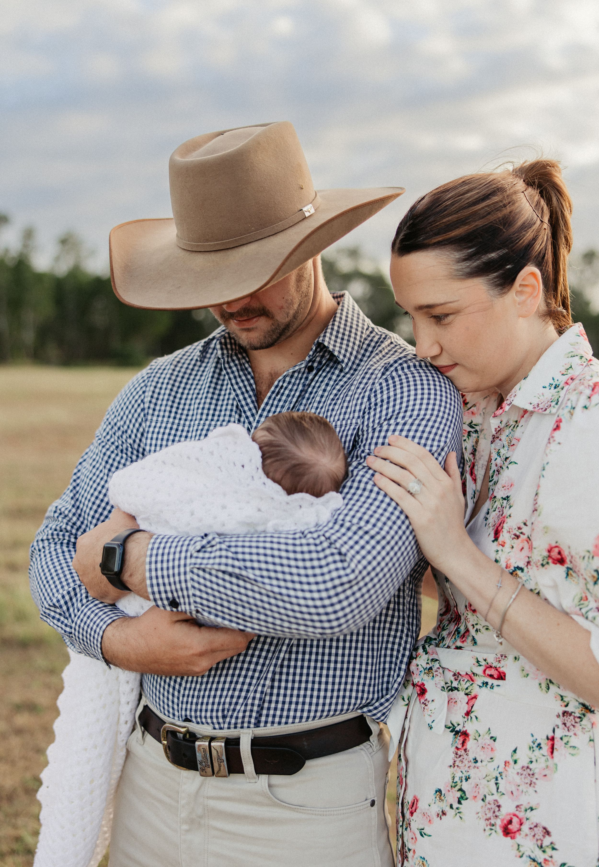 A couple in a field tenderly cradles their newborn baby, the man wearing a cowboy hat and blue gingham shirt.
