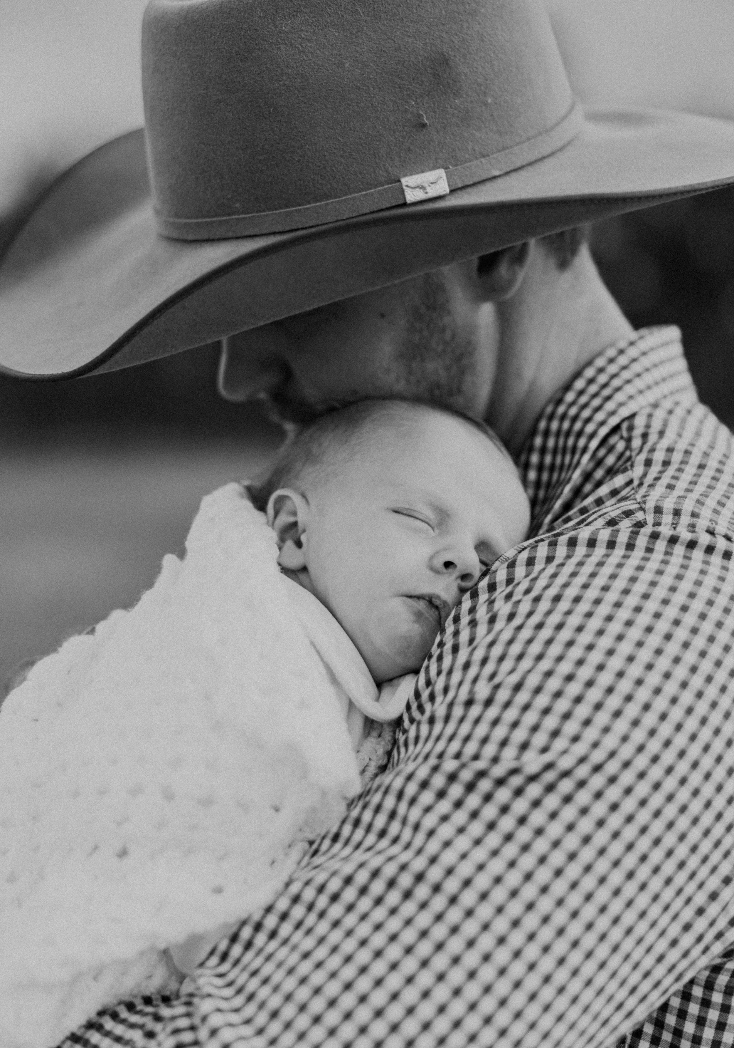 Black and white photo of a cowboy father in a wide-brim hat kissing his sleeping newborn baby wrapped in a white blanket.