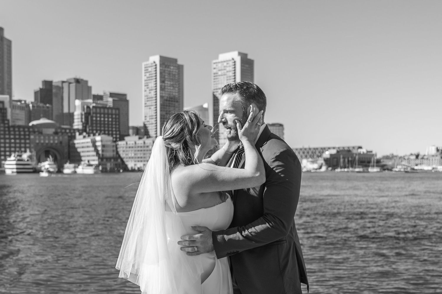 Black and white photo of a bride and groom embracing by the waterfront with a city skyline in the background.