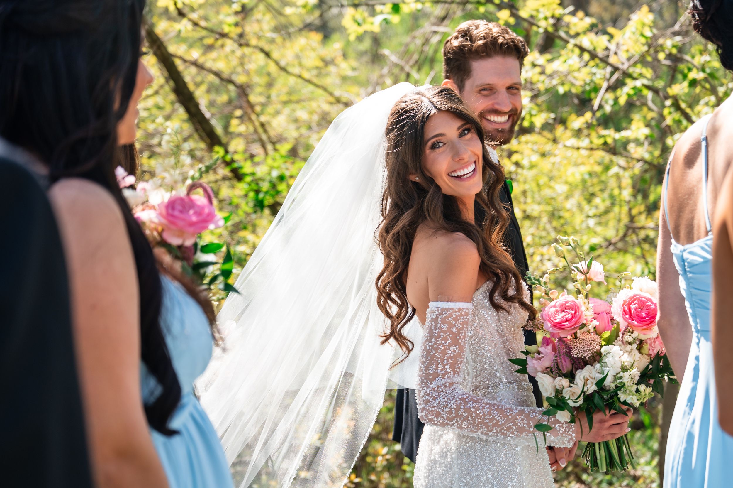 Bride in lace off-shoulder gown holding pink bouquet embraces groom outdoors with bridesmaids in blue dresses.