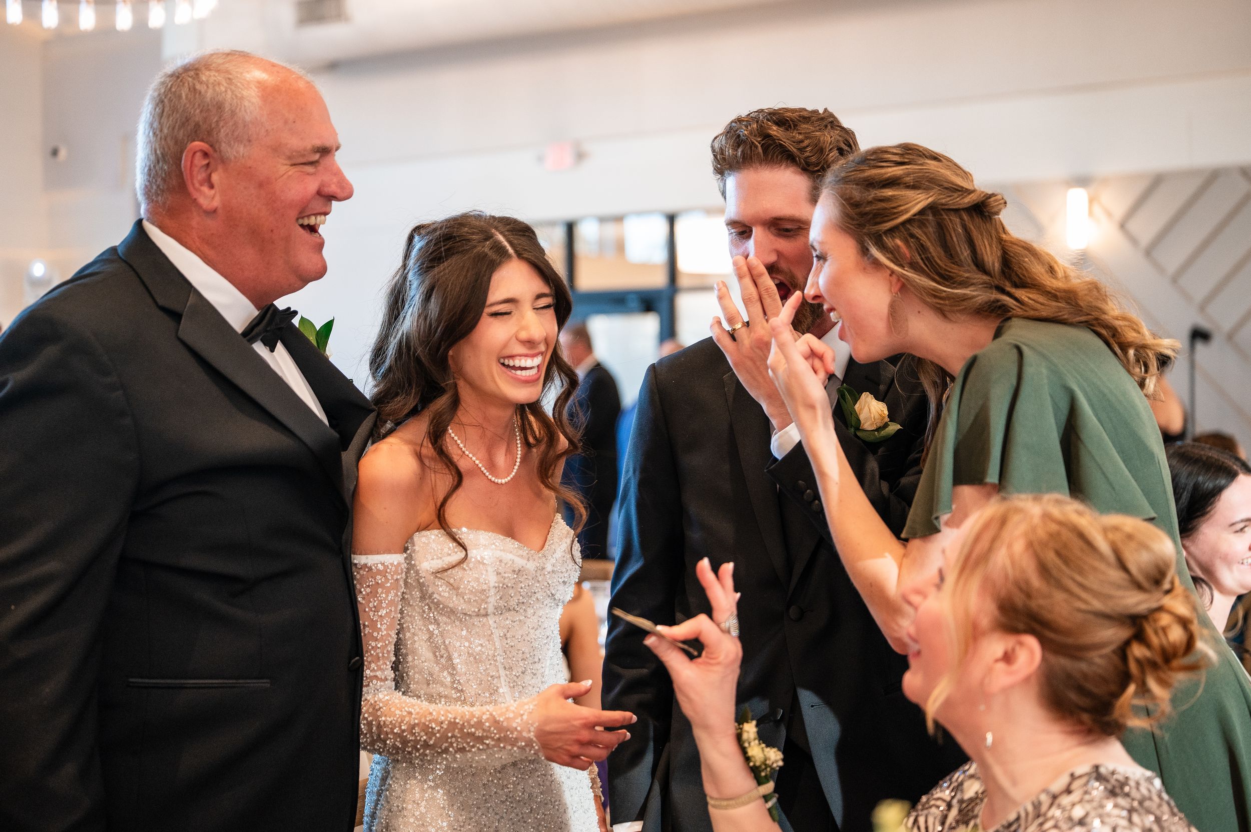 Bride in lace gown laughing with groom and wedding guests at a joyful indoor wedding reception celebration.