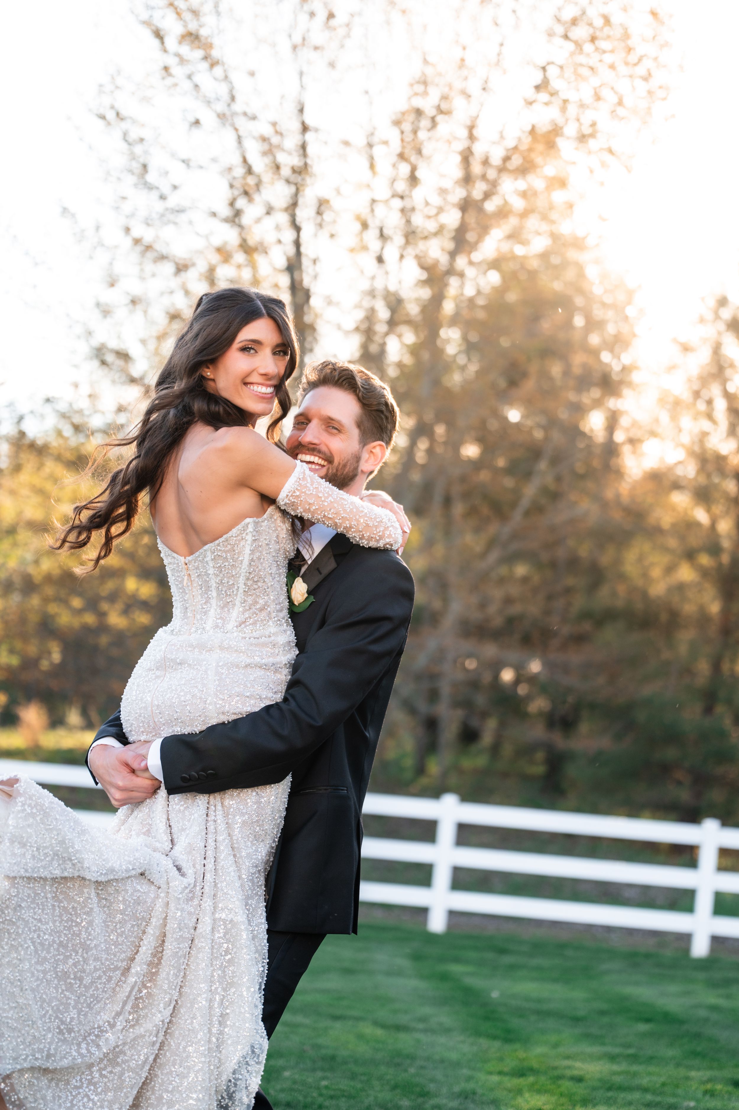 Bride in beaded gown held by groom in tuxedo, smiling outdoors at golden hour near white fence.