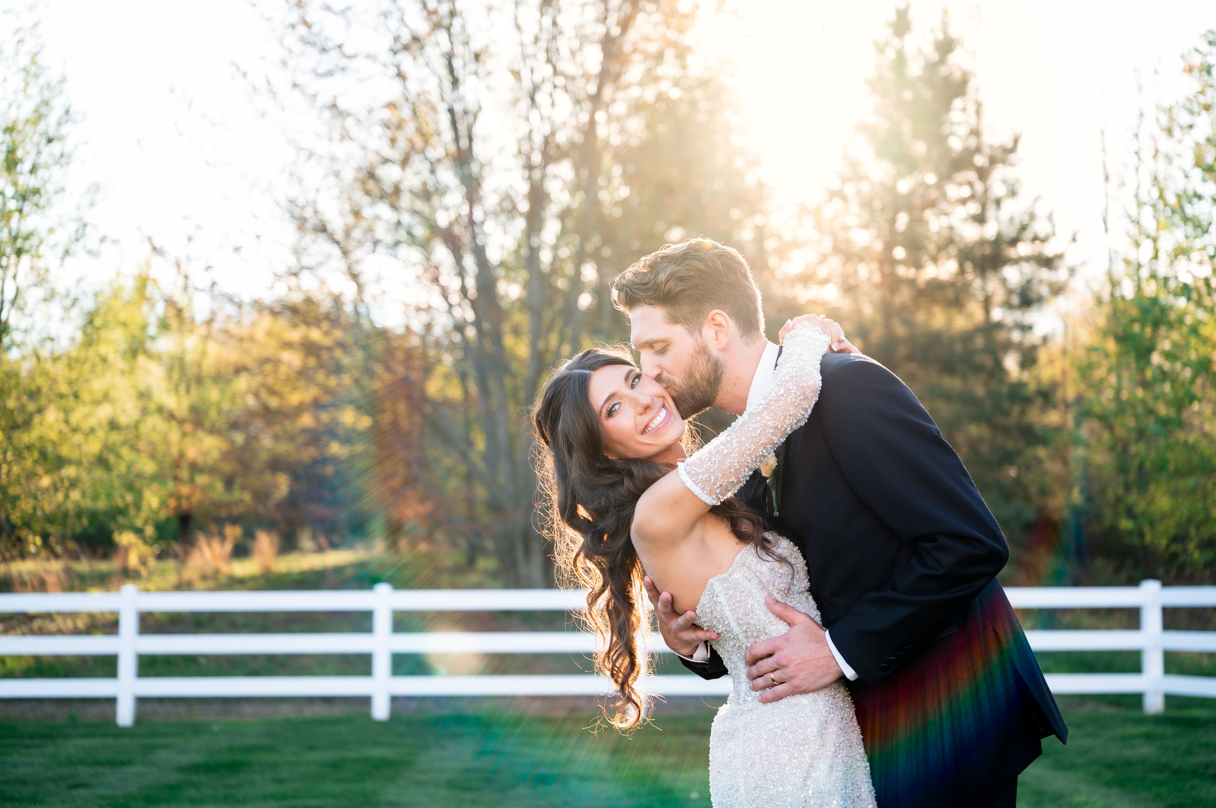 Couple embracing in golden sunlight at outdoor wedding venue with white fence and autumn trees.