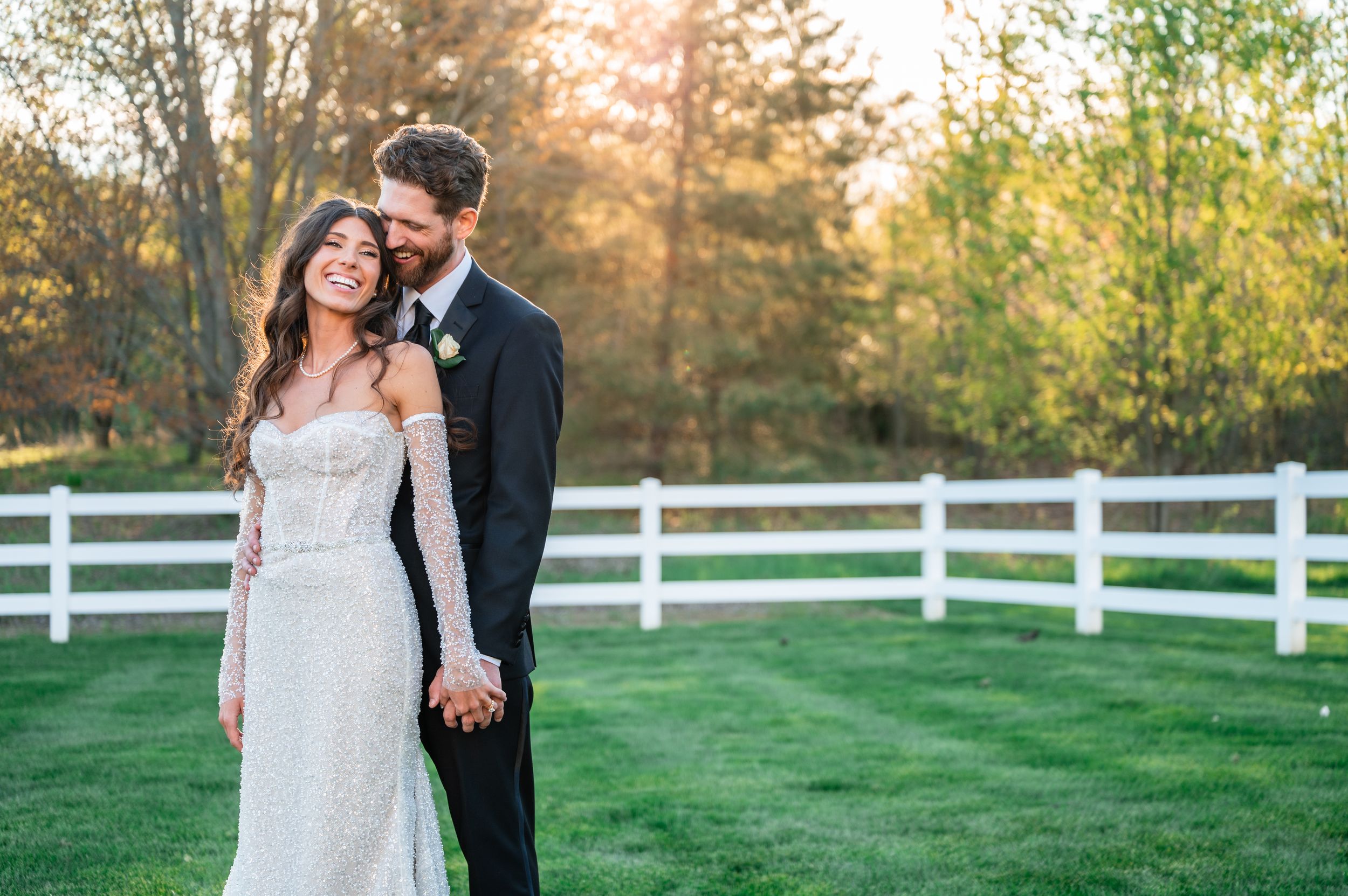 Bride and groom holding hands smiling outdoors at golden hour near white fence at rustic wedding venue.