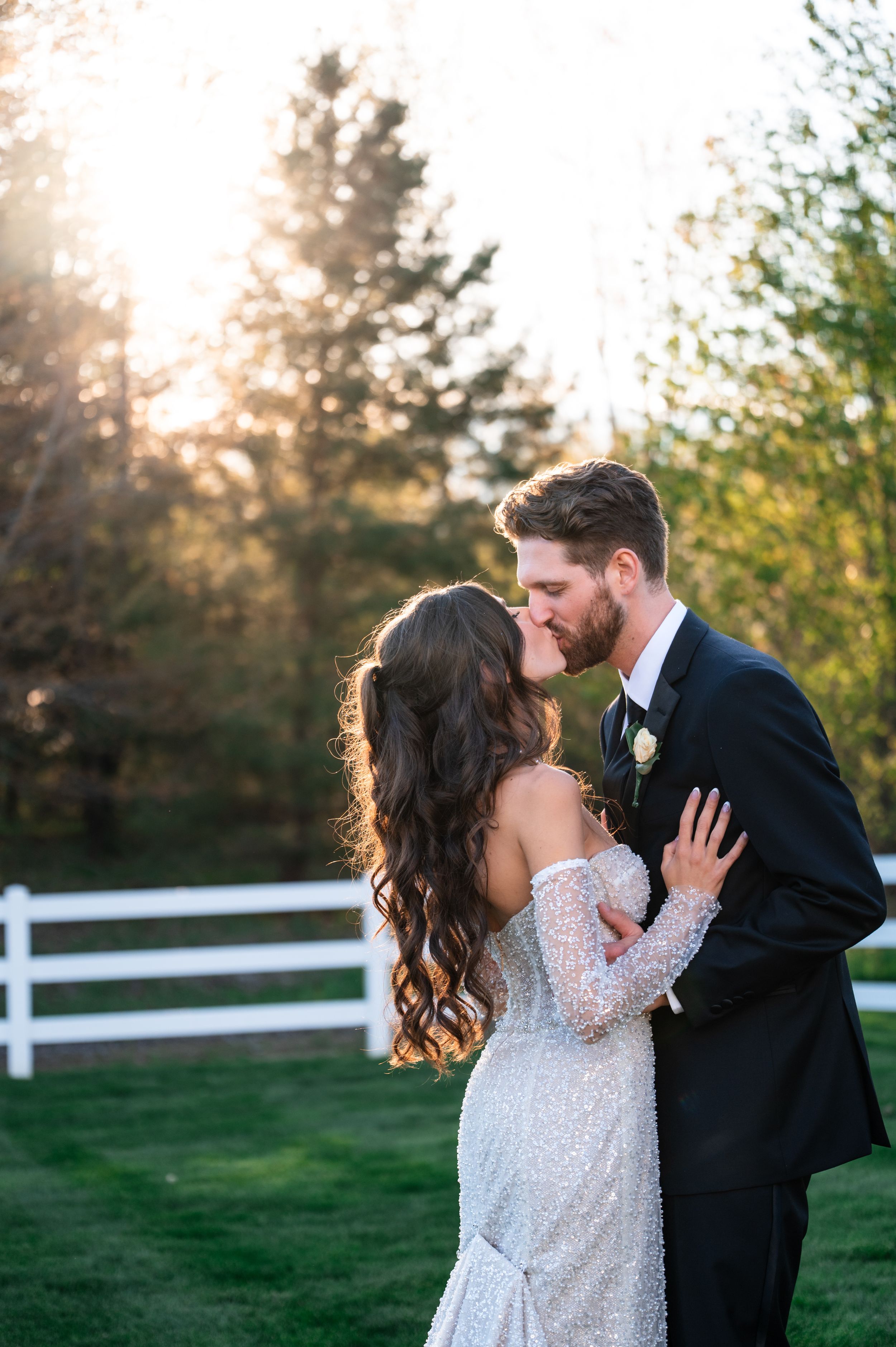 Bride and groom share a romantic kiss at golden hour, with a white fence and lush trees in the background.