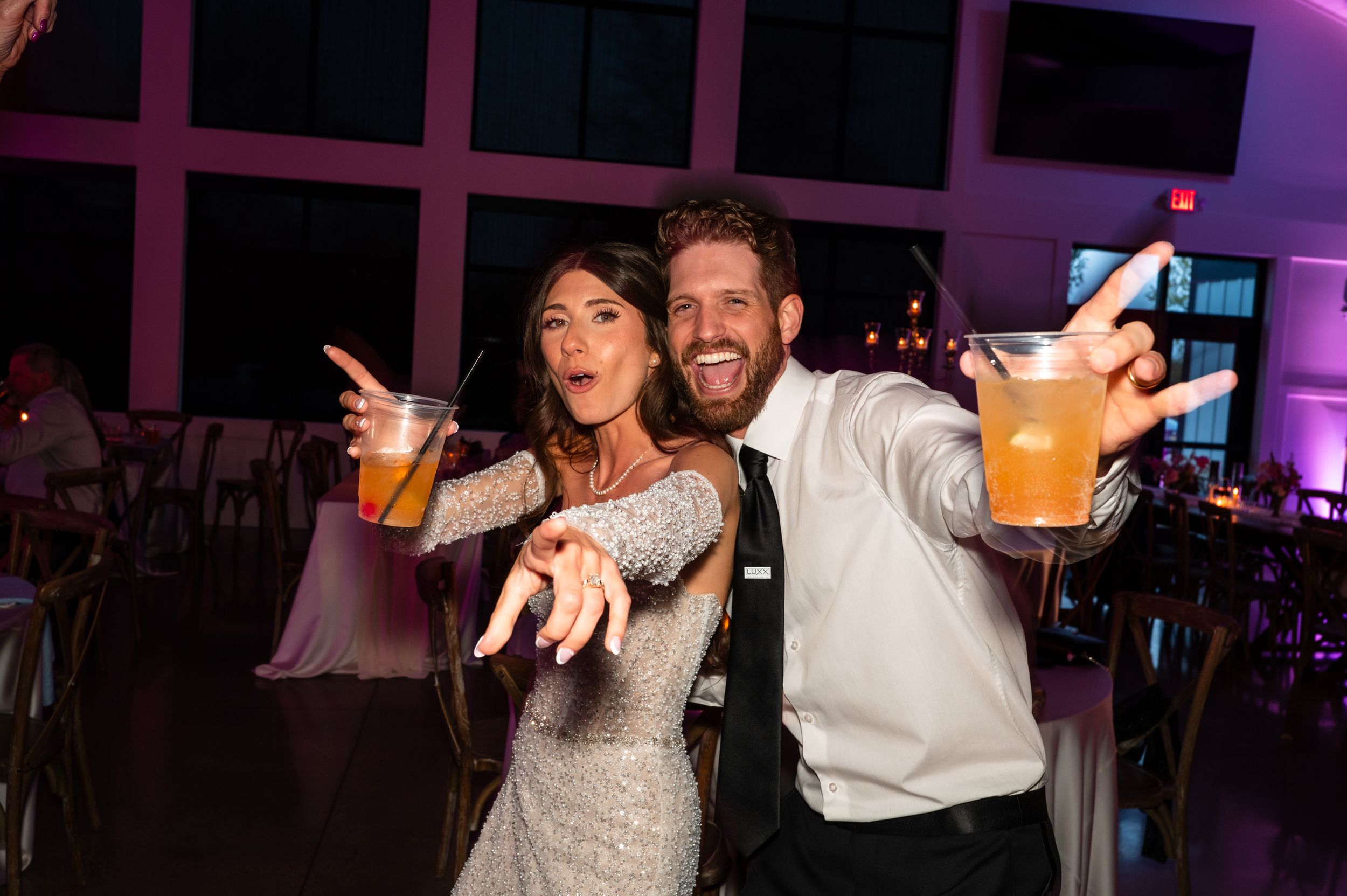Couple in formal attire toasting drinks at a wedding reception with pink ambient lighting.