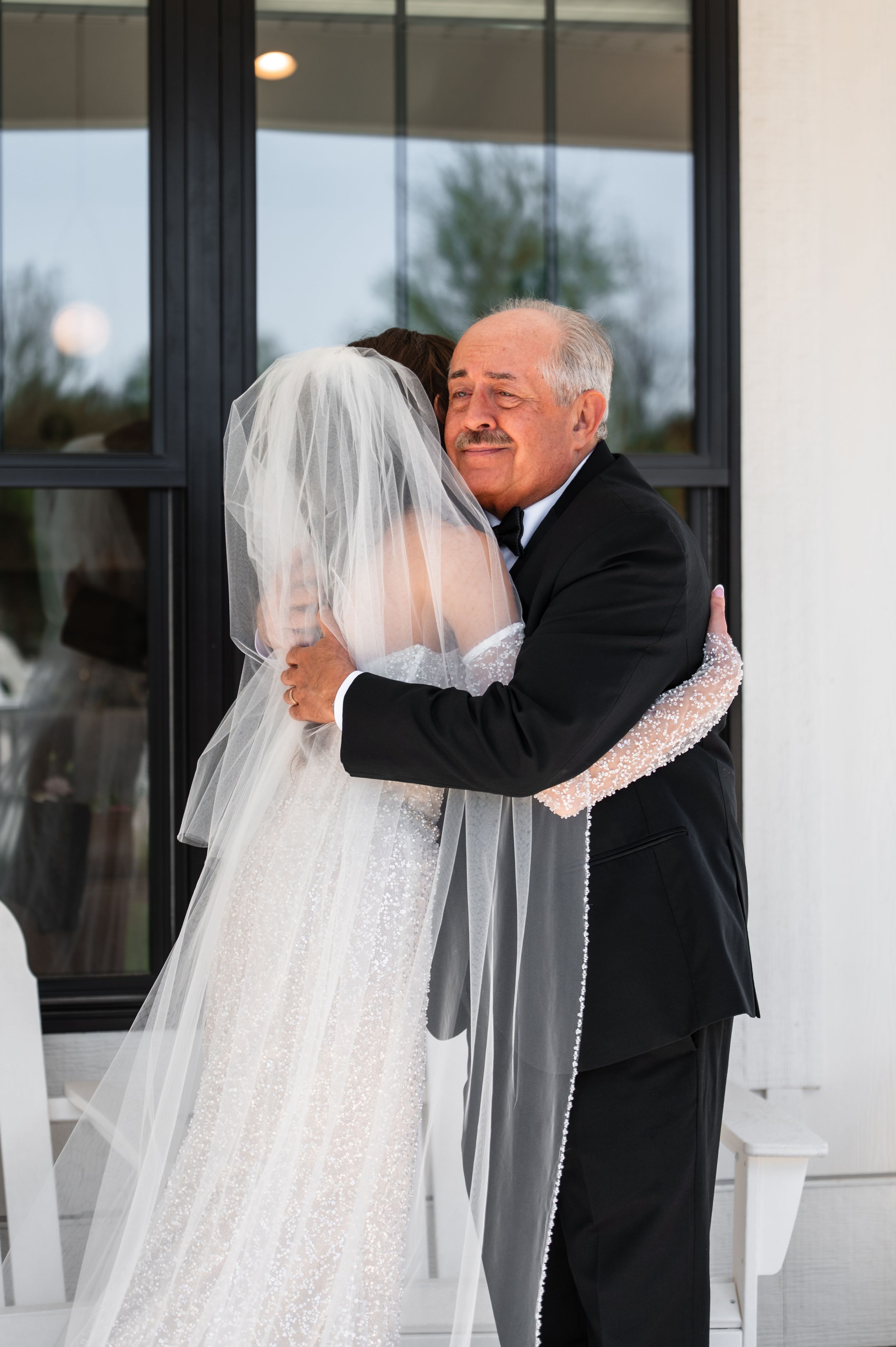 Emotional father-daughter wedding hug, bride in white gown and veil embracing older man in black tuxedo outdoors.