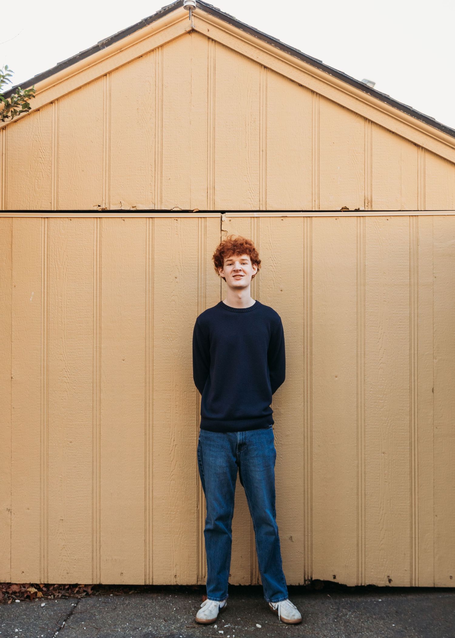 Young man with red curly hair in navy sweater and jeans smiling against a yellow wooden shed.