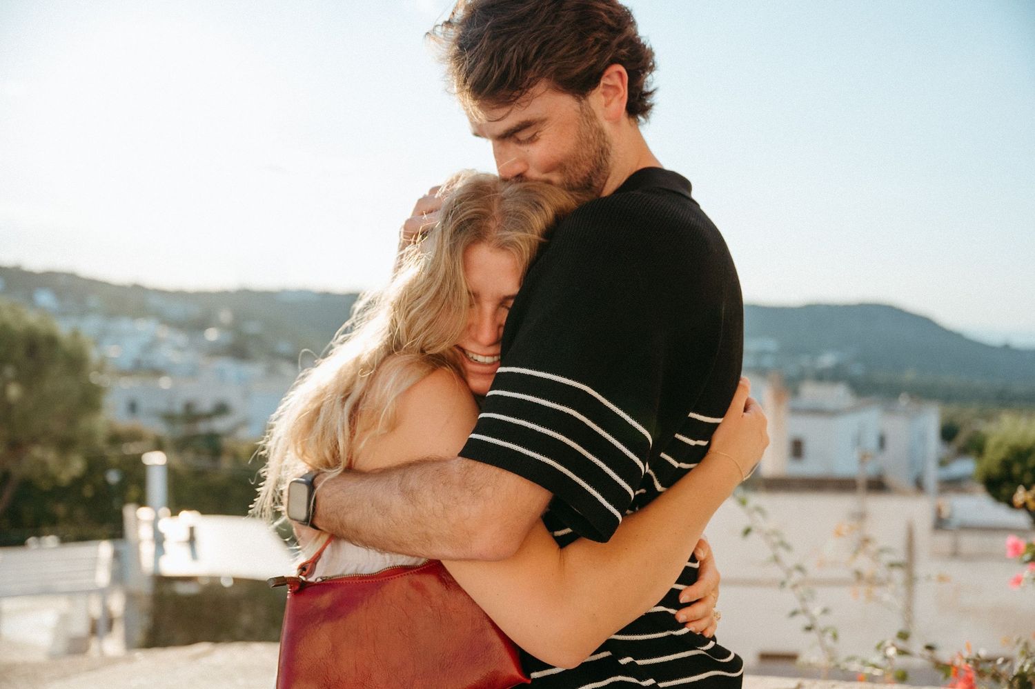 Young couple sharing a tender embrace outdoors with scenic Mediterranean village and sunny blue sky in background.