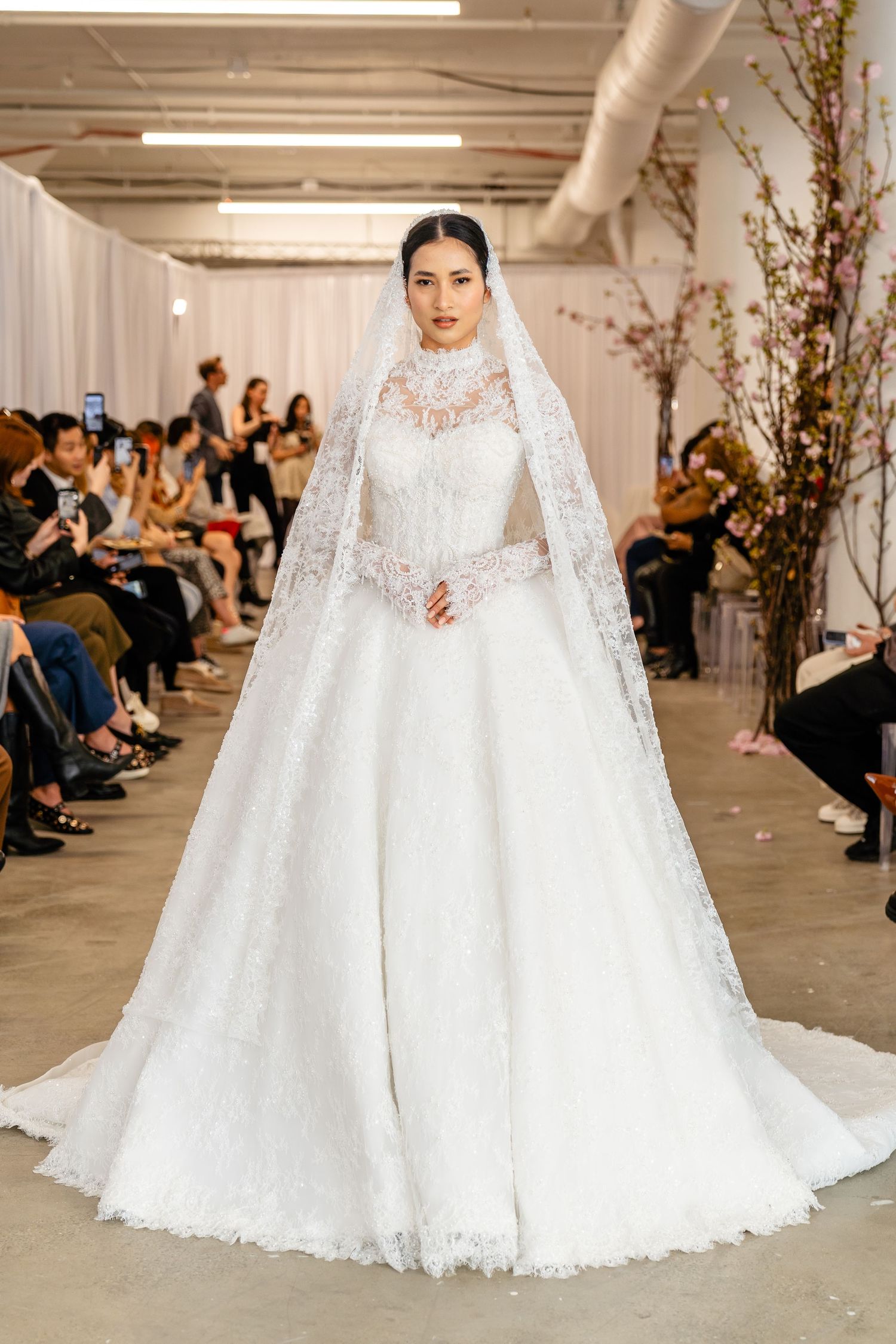 Model wearing an elegant white lace ball gown wedding dress with high neck and matching veil on runway.