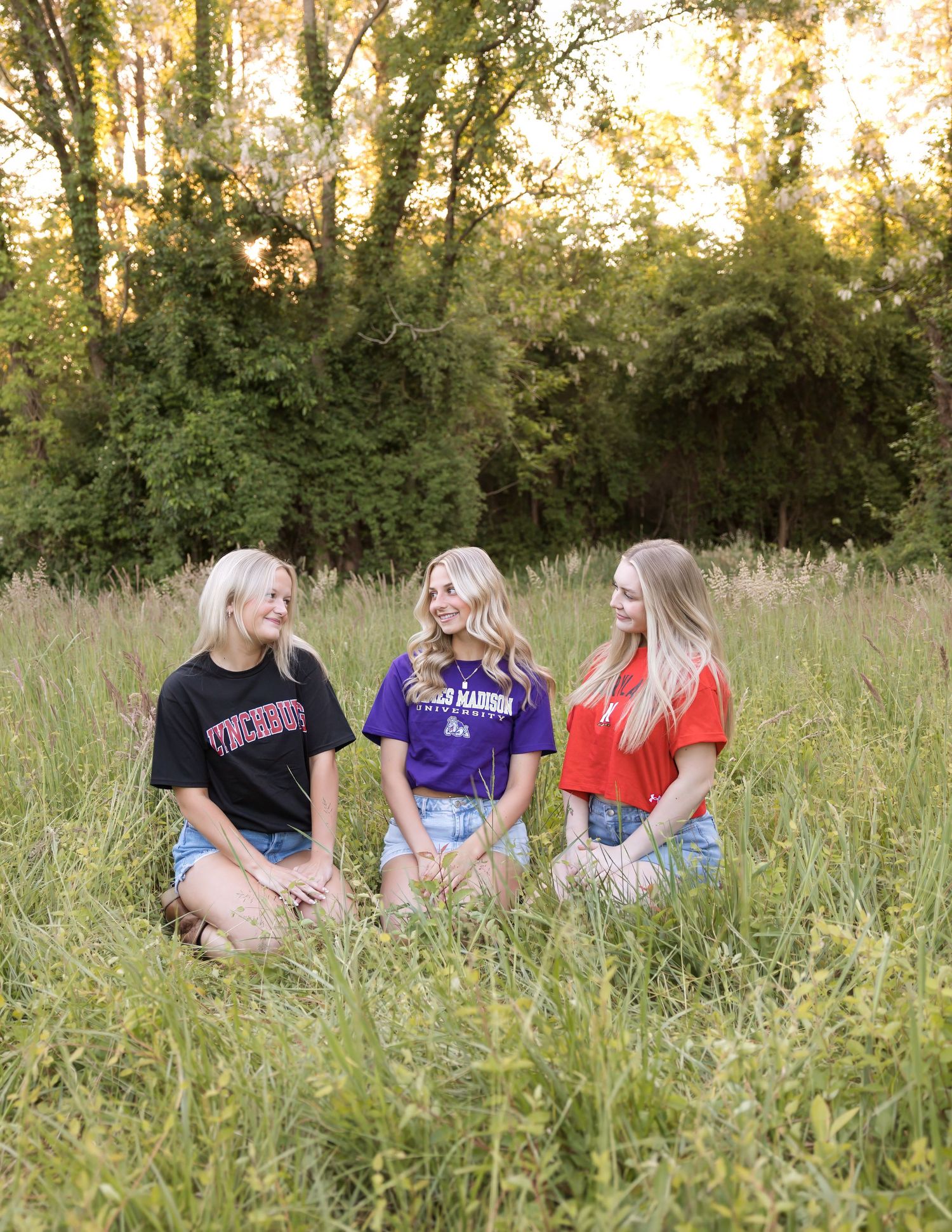 Three blonde women in white dresses sit together on a sandy beach watching a golden sunset over calm waters.