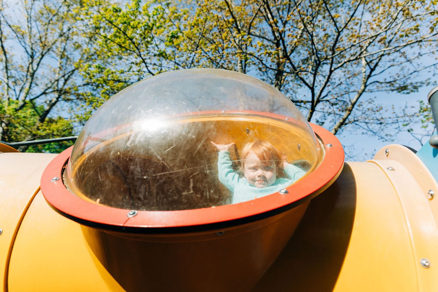 Young child waving from inside a clear dome bubble on a yellow playground structure outdoors.