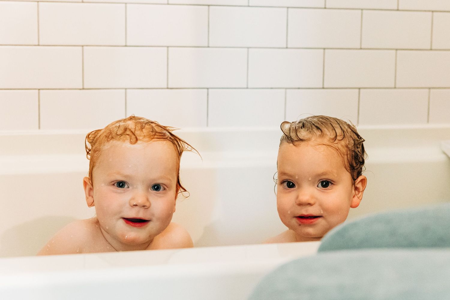Two smiling toddlers with wet hair sitting together in a white bathtub during bath time.