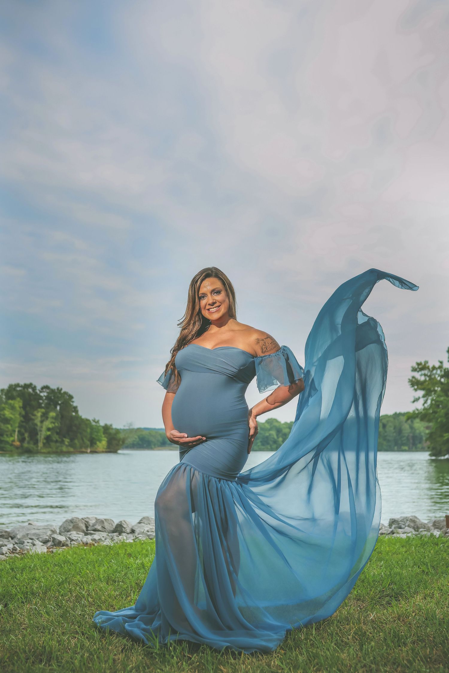 Pregnant woman in flowing blue off-shoulder gown poses by a lake with fabric billowing in the wind.