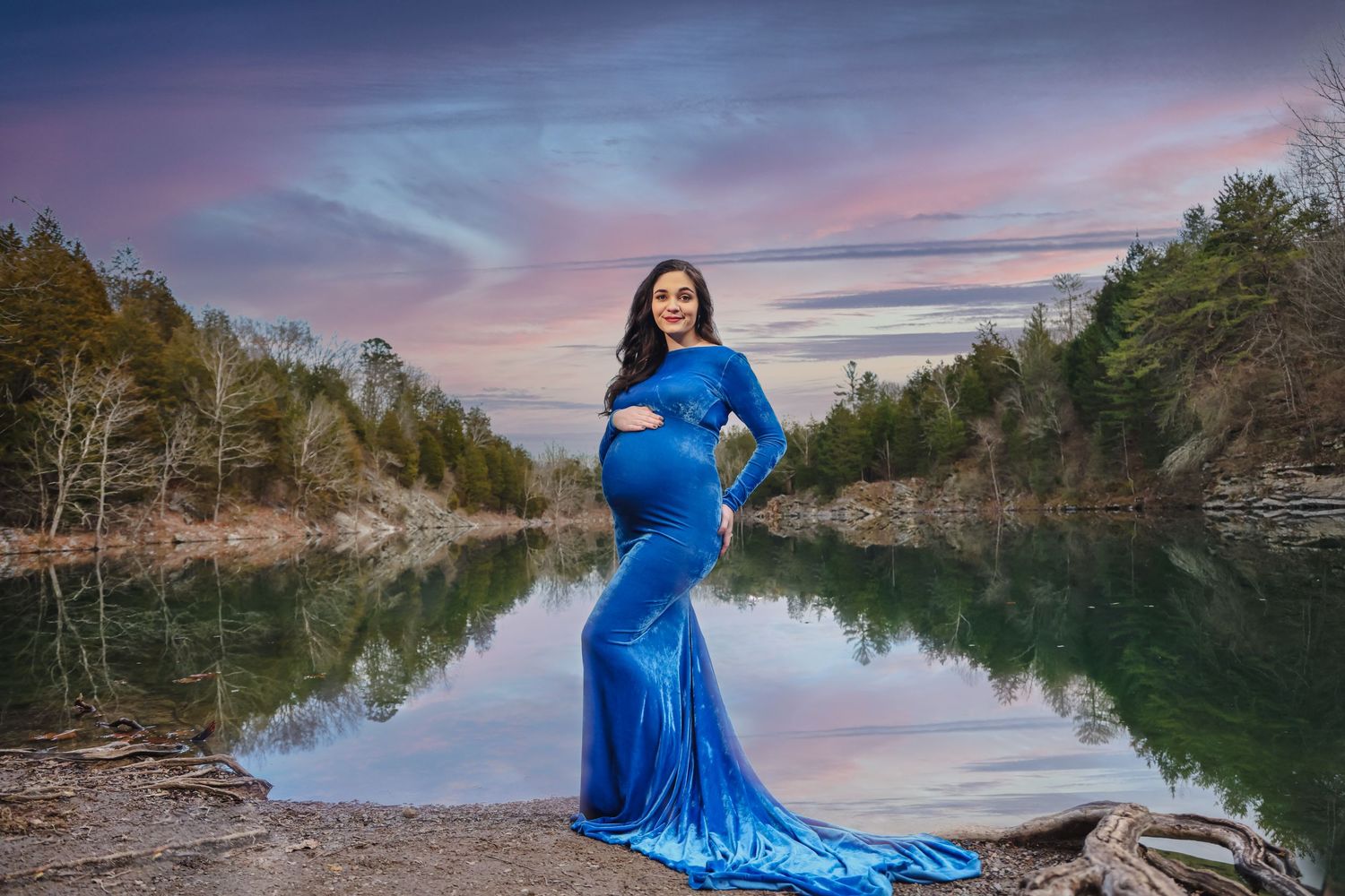 Pregnant woman in blue maternity gown poses by reflective lake at sunset with trees in background.
