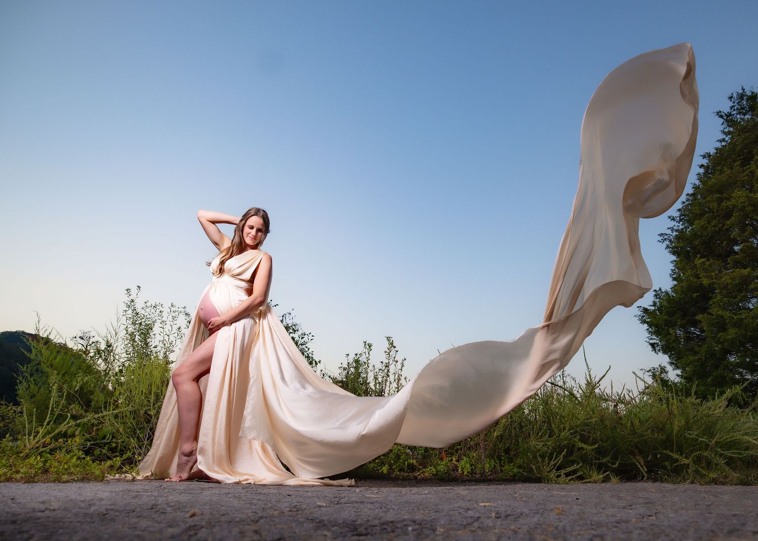 Pregnant woman in flowing champagne gown poses gracefully outdoors at sunset.