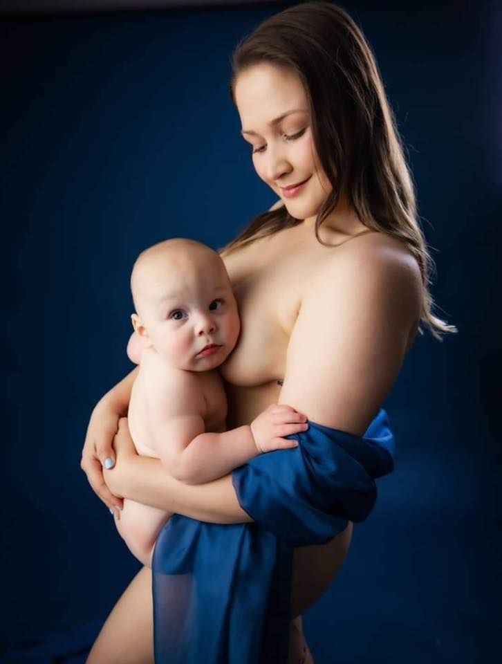 A mother tenderly holds her newborn baby wrapped in blue fabric against a dark blue studio background.