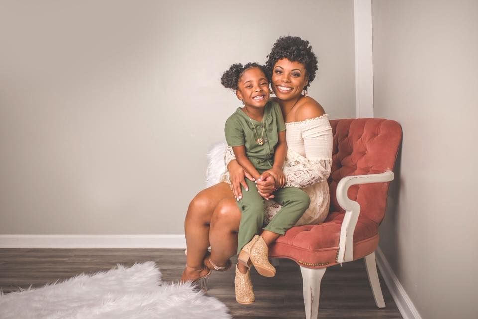 A mother and young daughter share a warm embrace on a pink velvet chair during a professional portrait session.